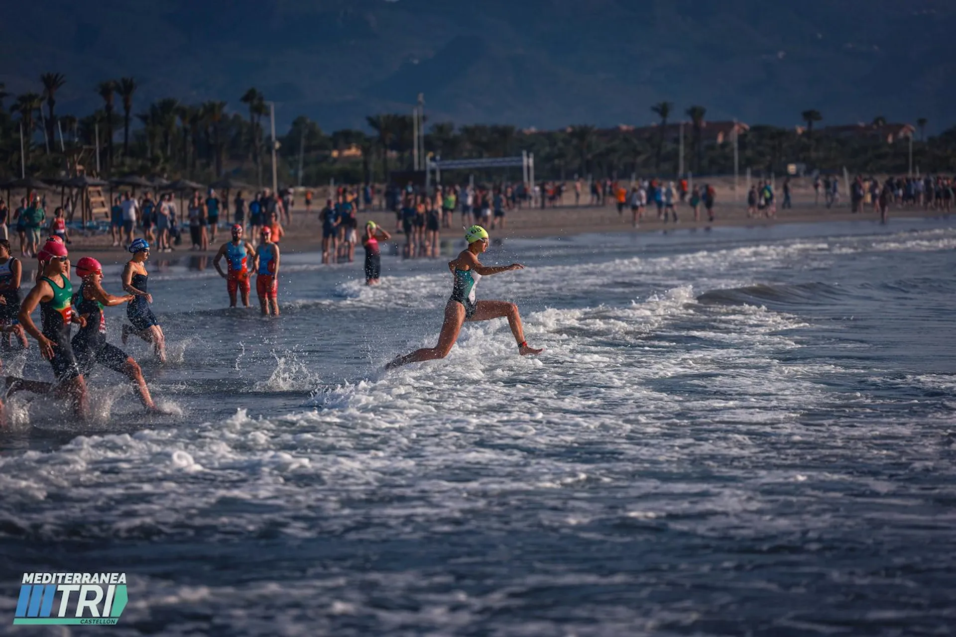 This image shows a group of athletes running into the ocean as part of a triathlon event. They're wearing swimsuits and swim caps, indicating the swim segment of the race. The beach is lined with spectators, and there are palm trees in the background. The "MEDITERRANEA TRI" logo suggests that it's part of the Mediterranea Tri event.
