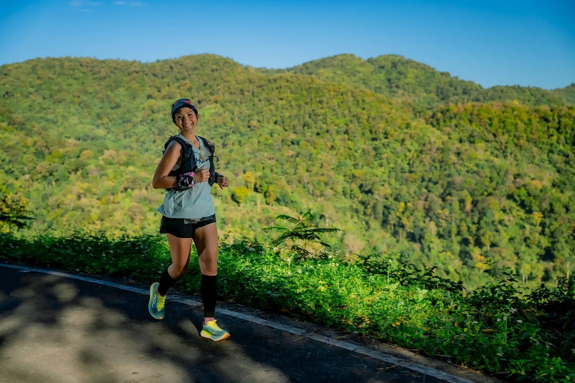 The image shows a person running outdoors on a paved path. They are wearing a tank top, shorts, a cap, sunglasses, and running gear. The background consists of green, forested hills and a clear blue sky.