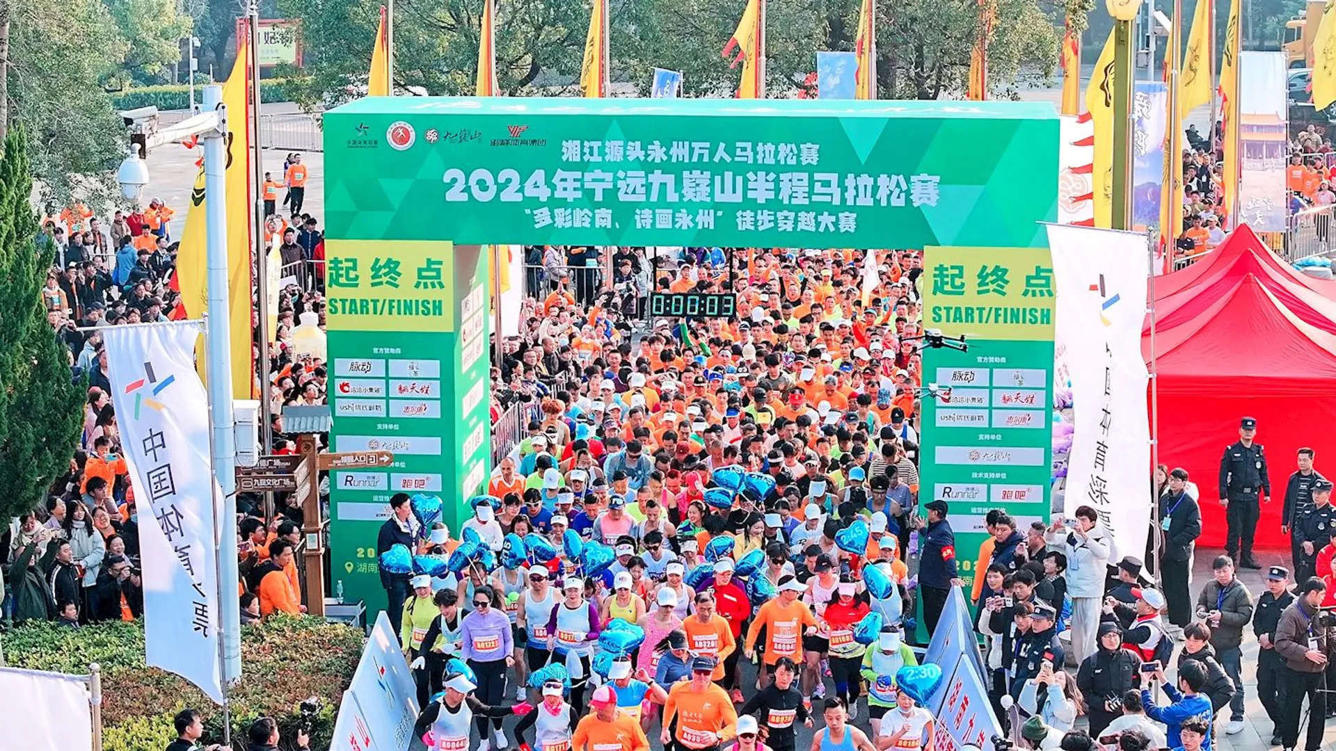 The image shows the starting line of a marathon or running event. There is a large group of runners waiting to begin the race. A banner above the crowd displays information about the event, including "2024" and "marathon." There are also flags and banners, as well as event staff around the area. The place appears crowded and lively.