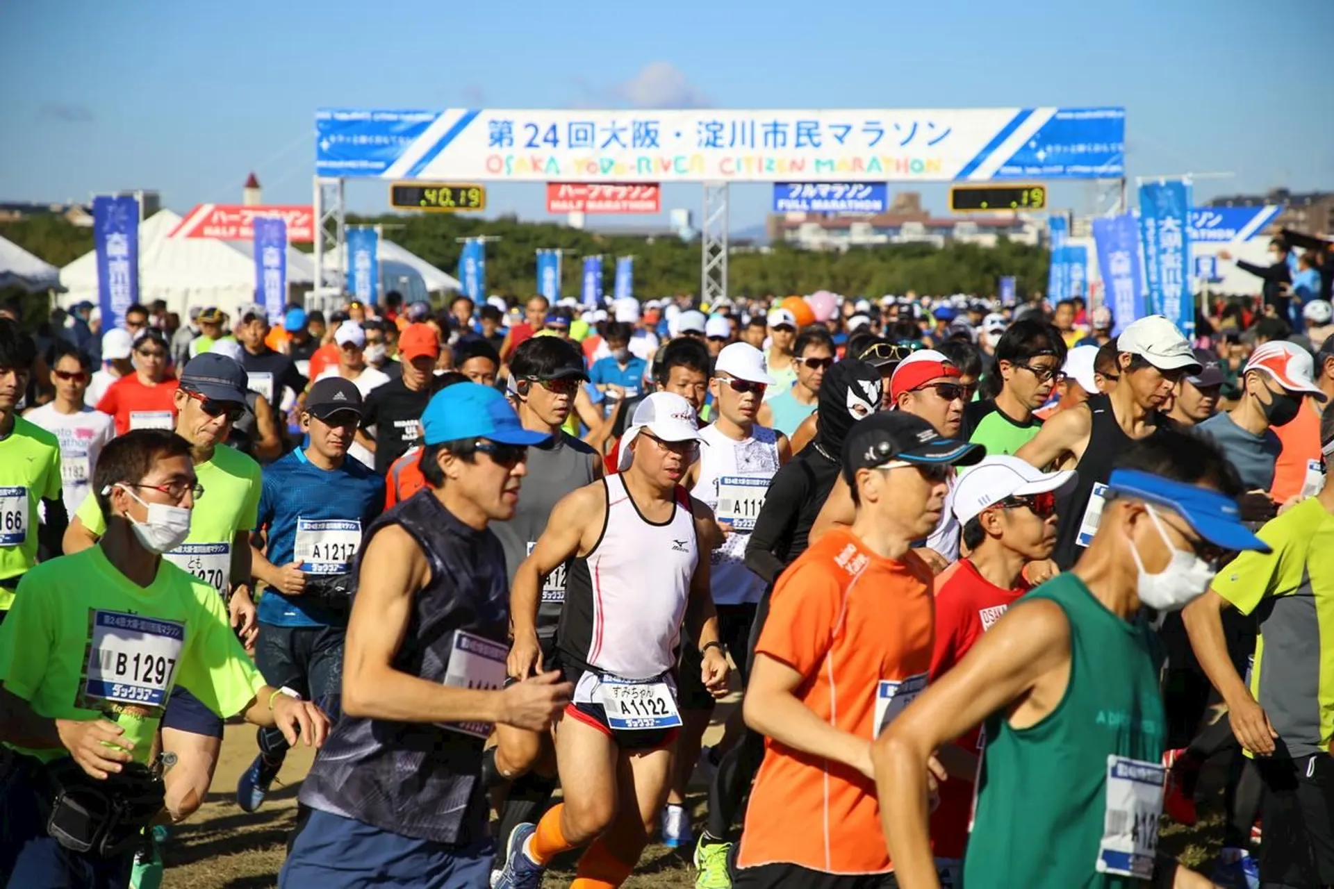 The image shows a large group of people participating in a running event, possibly a marathon or other long-distance race. The participants are dressed in athletic clothing suitable for running, with many wearing specialized running shoes, shorts, and t-shirts. Some runners have bibs on their fronts with numbers, which are common in organized races for identification and tracking purposes.

In the background, there is a start or finish line arch with banners and signs written in Japanese, indicating that the event is taking place in Japan or is related to Japanese culture. The banners are likely showing the name of the event, sponsors, and possibly the distance of the race. The sky is blue and clear, suggesting that the weather is favorable for an outdoor event. There is also a crowd