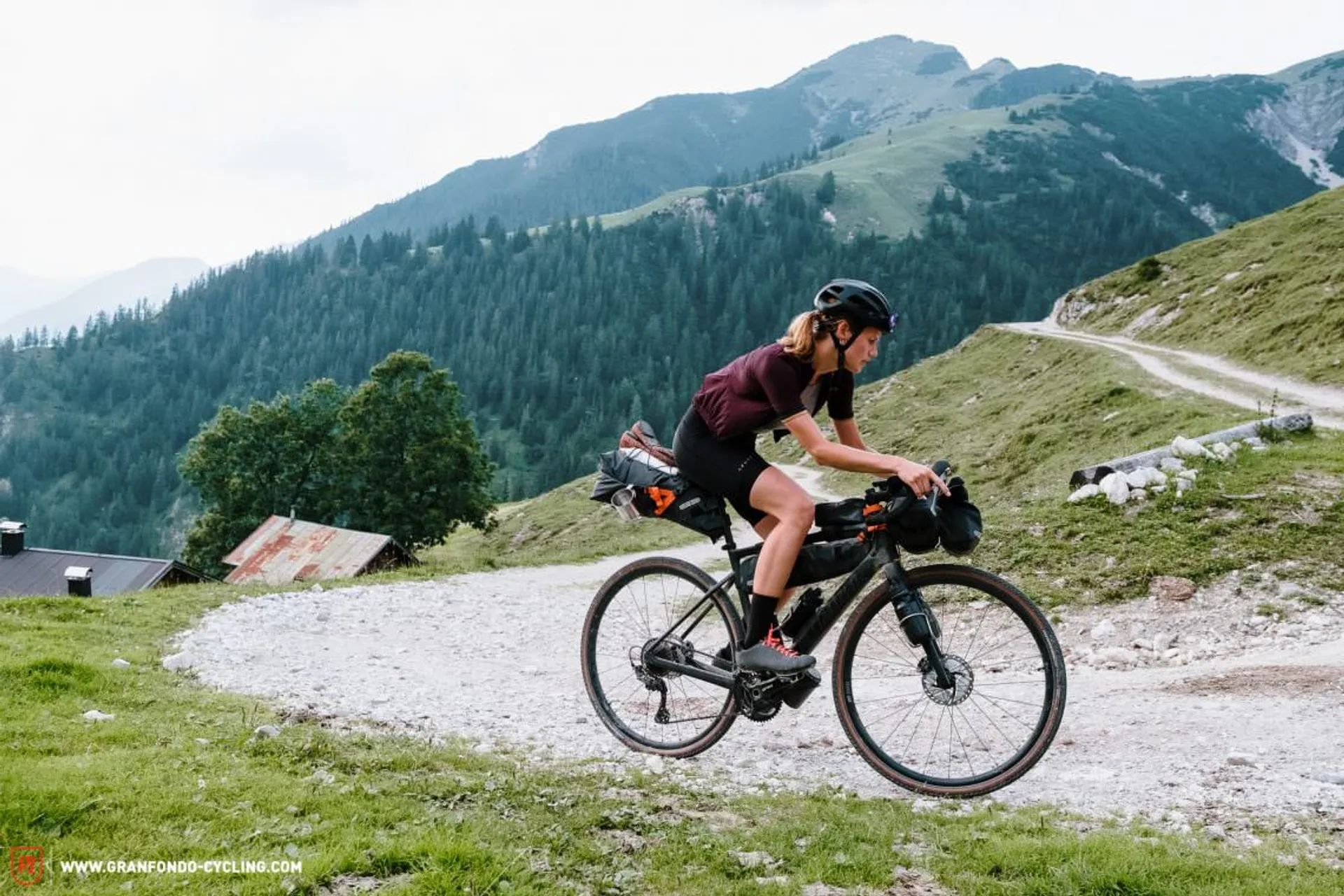 The image shows a cyclist riding on a gravel path in a mountainous landscape.