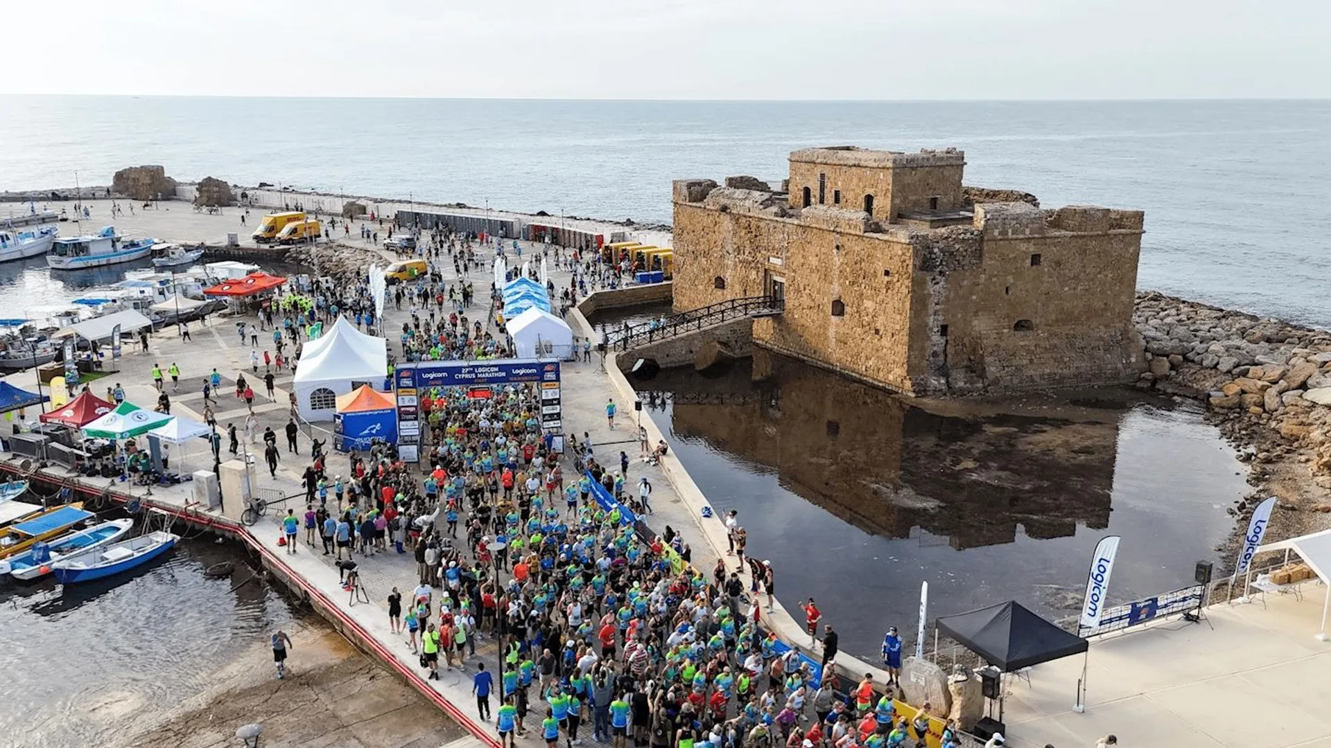 The image shows a large group of people gathered for what appears to be a running event near a historical fortress by the sea. There are tents, banners, and a starting line, suggesting the beginning of a race. The fortress is situated on a pier surrounded by water, and there are boats and vehicles in the area.