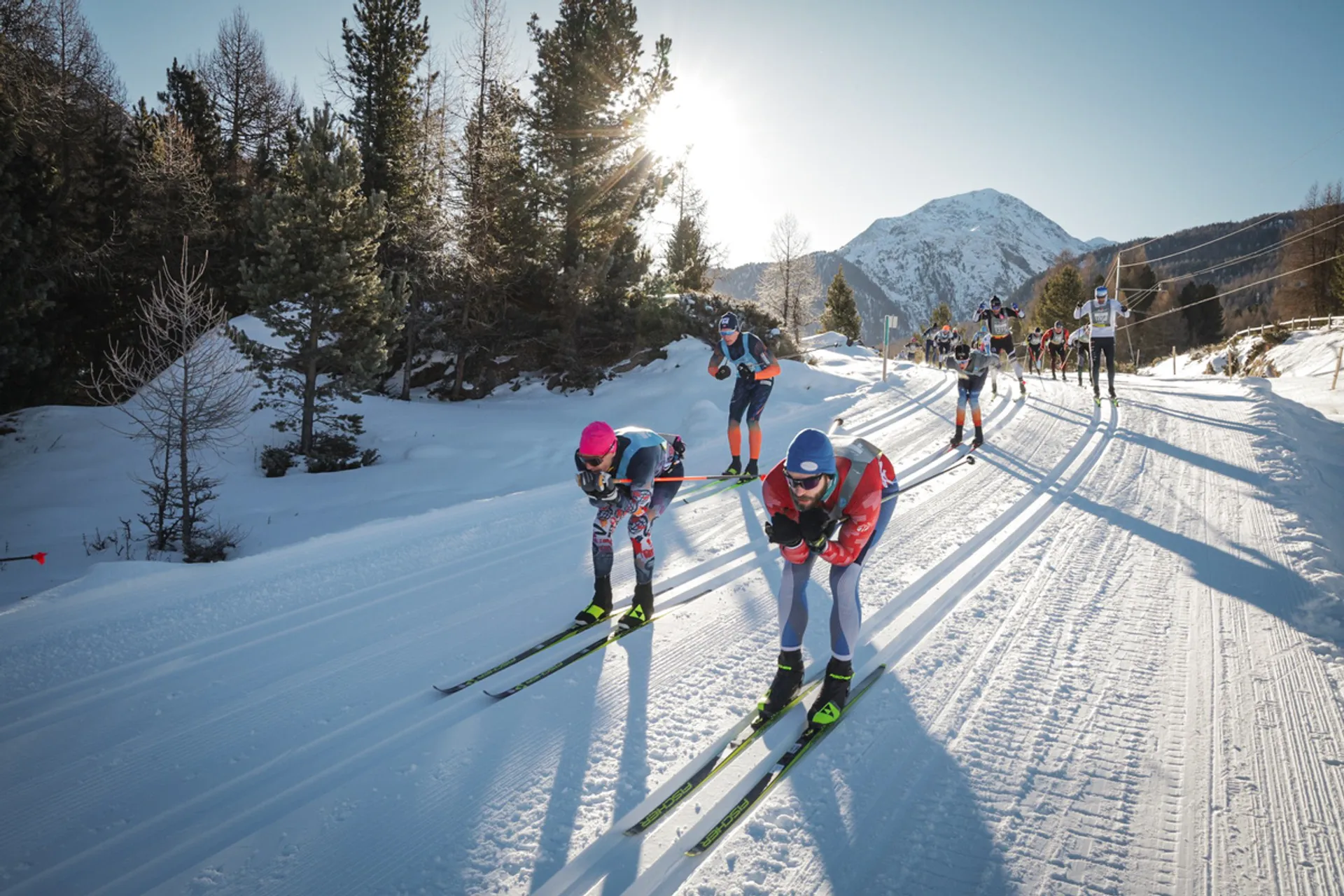 This image shows a group of people cross-country skiing on a snowy trail with a mountainous background. The skiers appear to be in a race or organized event. Sunlight is shining, creating a clear and bright atmosphere with trees lining the path.