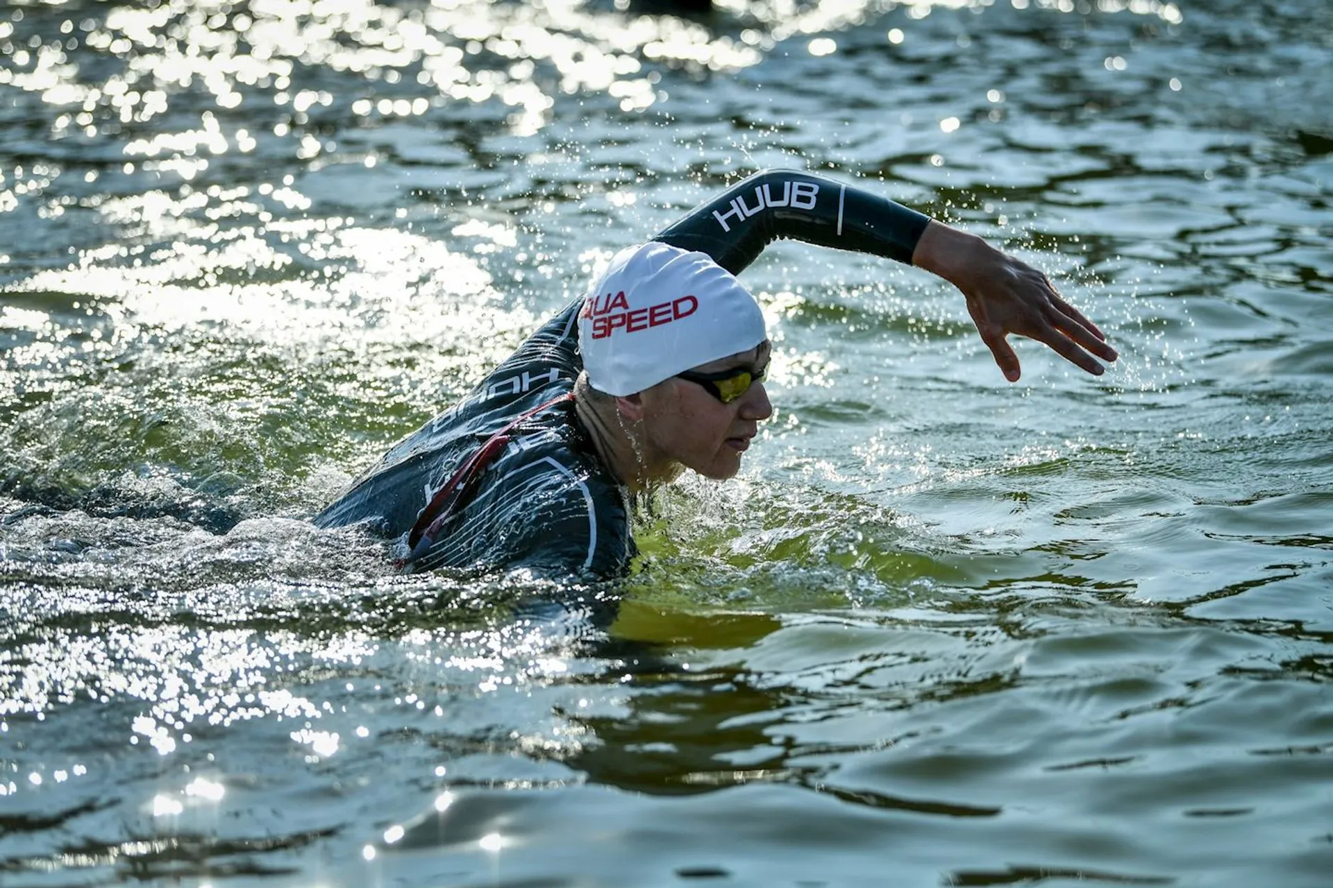 The image shows a person swimming in open water. They are wearing a wetsuit, swim cap, and goggles, and are performing a freestyle stroke. The lighting suggests it might be during the day with sunlight reflecting off the water.