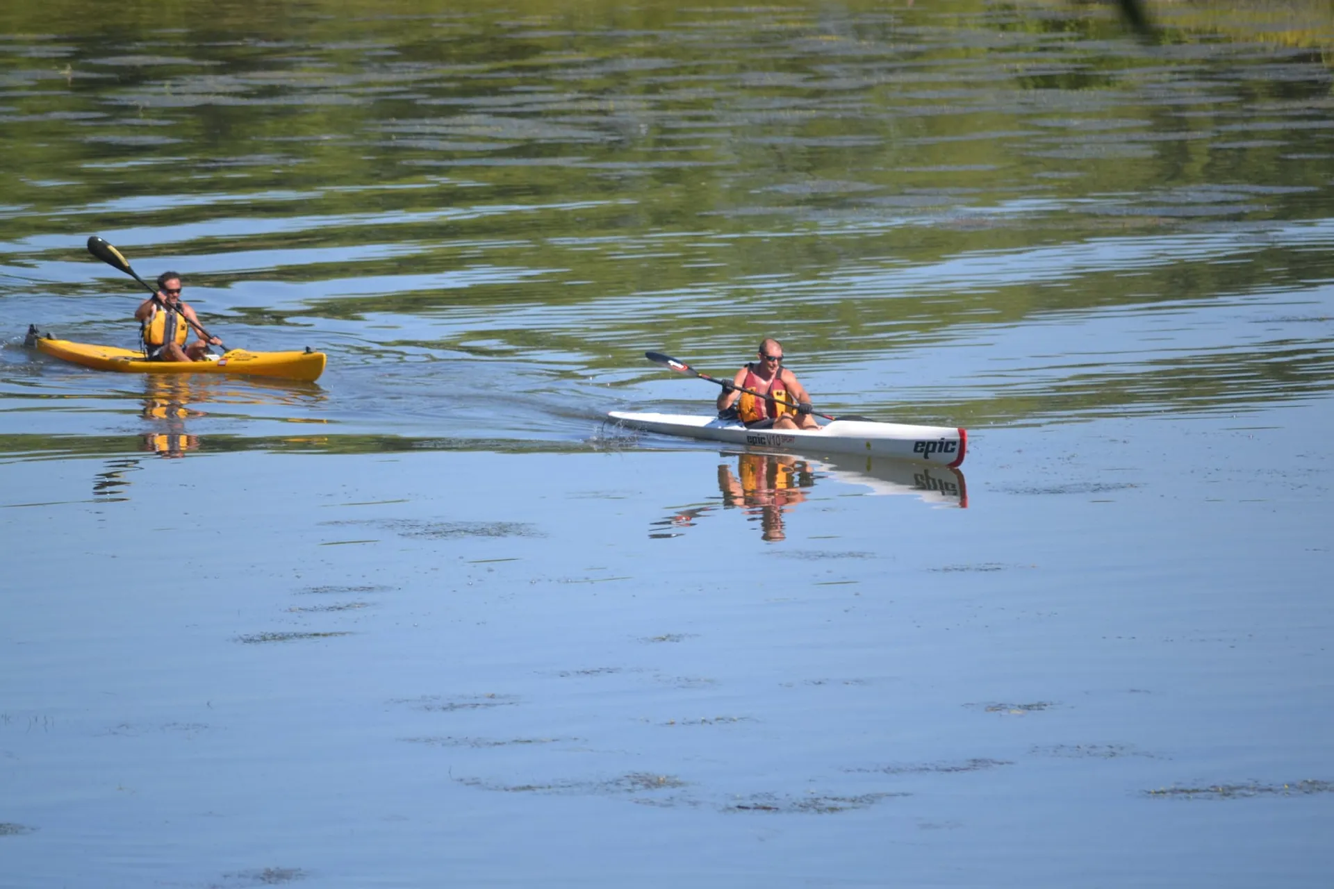 The image shows two individuals kayaking on a body of water. The person in the foreground is paddling a white kayak with the word "epic" visible on the side, indicating the kayak brand. The individual appears to be wearing a life vest for safety, and they are using a double-bladed paddle. The second person is farther back, in a yellow kayak, similarly equipped with a double-bladed paddle and a life vest. Both are actually using paddles appropriate for kayaking, showing that they are likely enjoying recreational time on the water. The water is calm, and there are reflections visible on the surface, suggesting a peaceful, serene environment. Vegetation can be seen on the banks in the background, indicating that this scene is likely