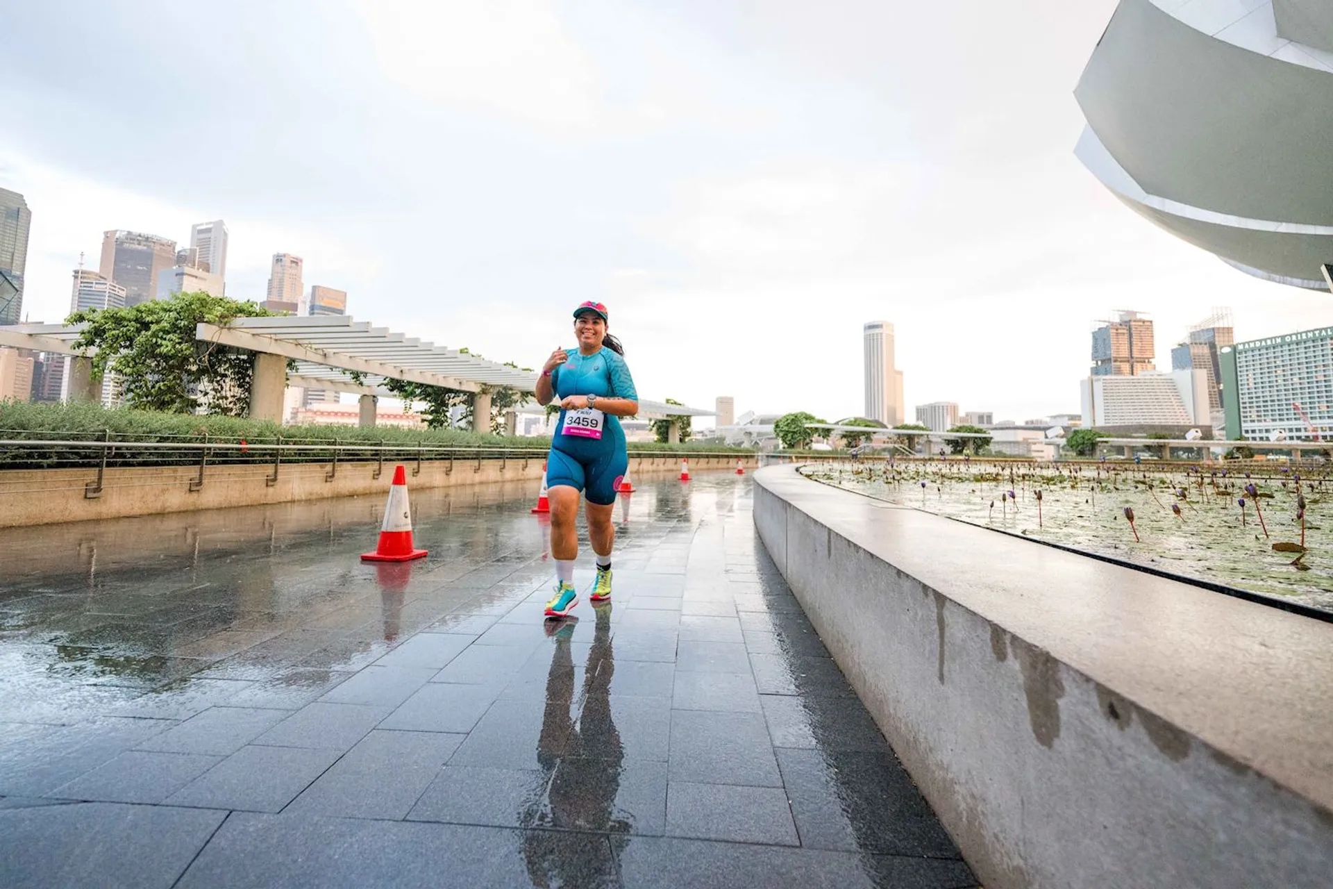 The image shows a person running on a wet pathway in an urban area. There are skyscrapers in the background, and some cones placed along the path. The person is wearing athletic gear, including a cap and a race bib. The environment appears modern with some greenery and architectural elements nearby.