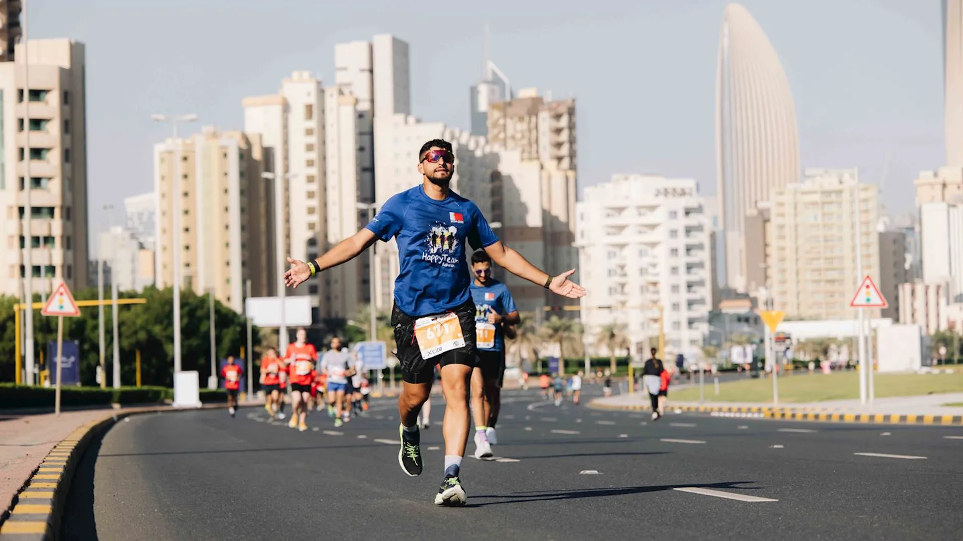The image shows a person running in a race on a city street with skyscrapers in the background. The runner is wearing sunglasses, a blue shirt with a race bib, and is gesturing with open arms. Other participants can be seen behind him. The street is marked for traffic, and warning signs are visible along the road. It appears to be a sunny day.