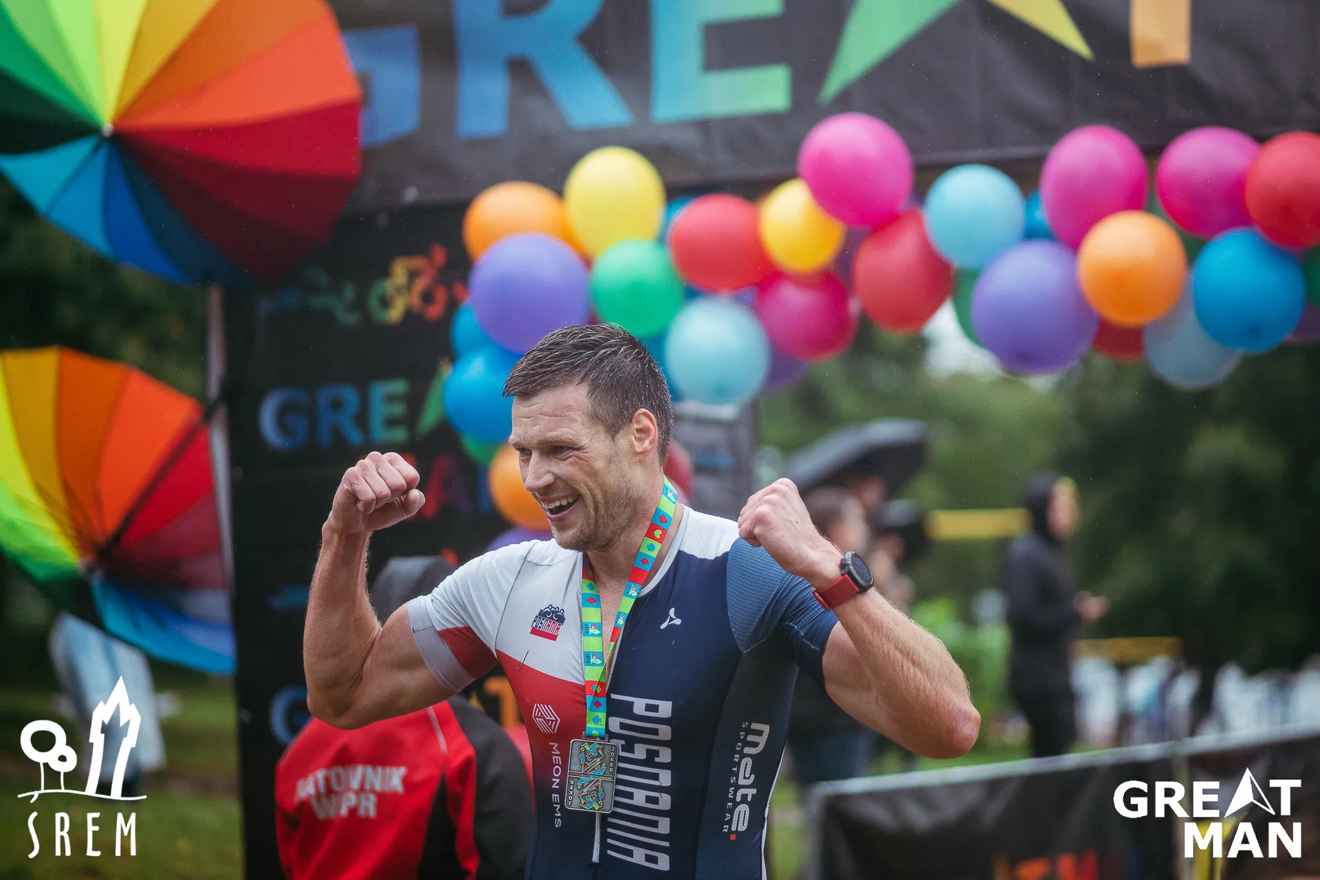 The image shows a person celebrating at a finish line of a sports event. They are wearing a race suit with the word "Poznań" on it and have a medal around their neck. There are colorful balloons and banners in the background.