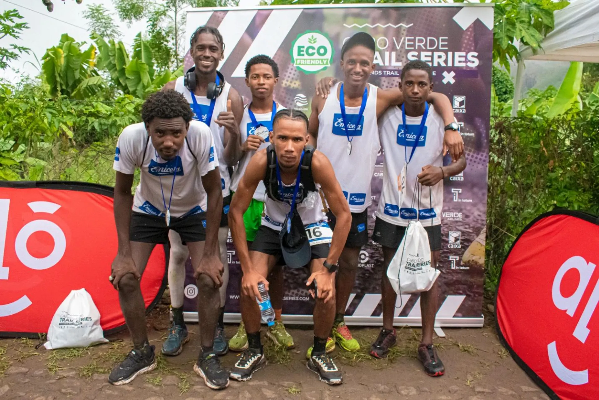 The image shows a group of six individuals posing together after what appears to be a trail running event. They are standing in front of a backdrop that reads "Eco Verde Trail Series" and holding goodie bags. Some of them are wearing medals, indicating they participated in or completed the event. There are also sponsor logos on the backdrop and on the sides. The setting appears to be outdoors, with greenery in the background.