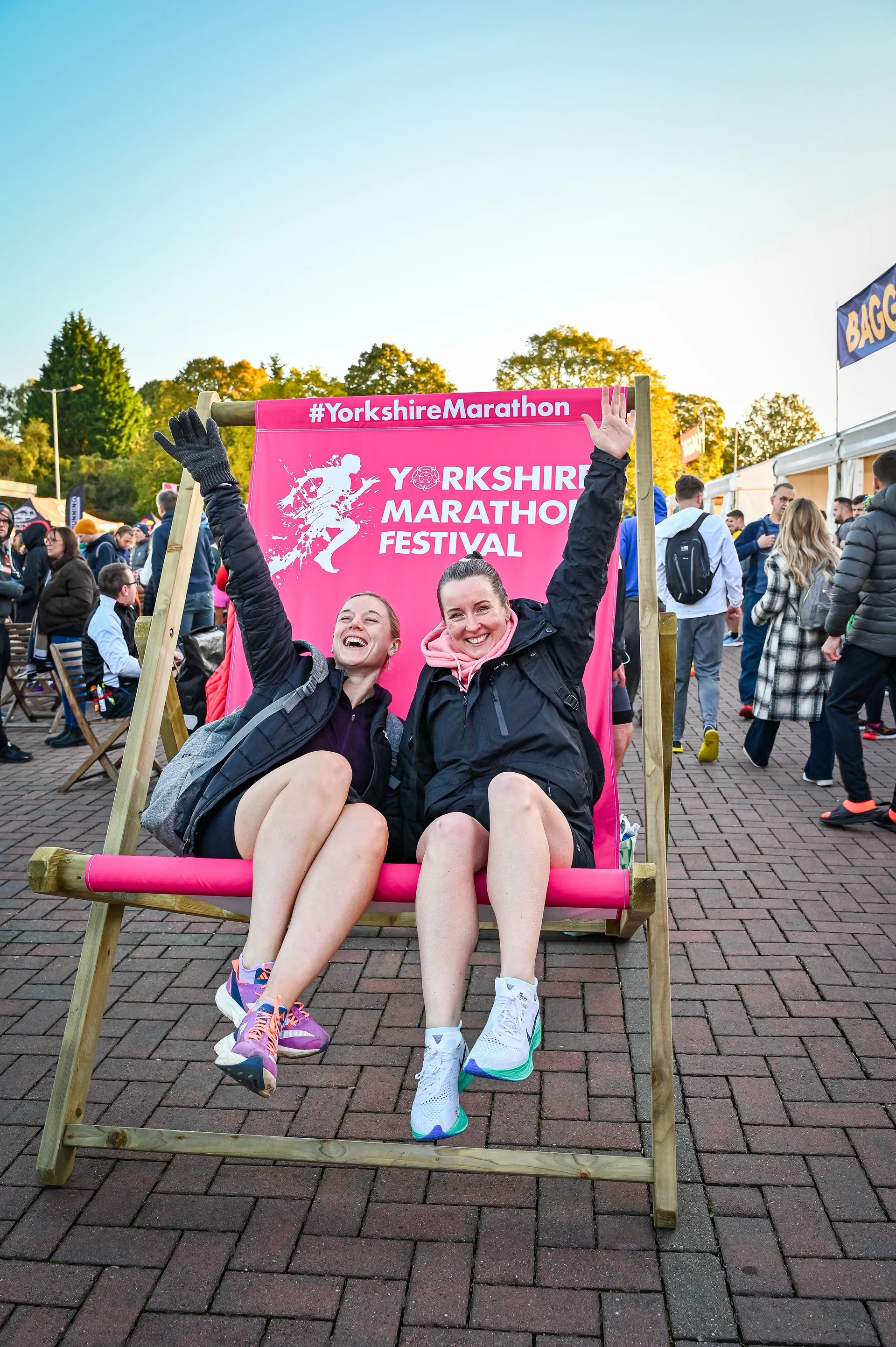 The image features two individuals sitting in an oversized deck chair, celebrating with their arms raised in excitement. They appear to be at the Yorkshire Marathon Festival, as indicated by the branding on the chair. Their joy could be related to participating in the event or simply enjoying the atmosphere at the festival. They are dressed in casual, sporty attire, indicative of a relaxed or athletic occasion, and there's a crowd of people in the background, suggesting a public outdoor setting.