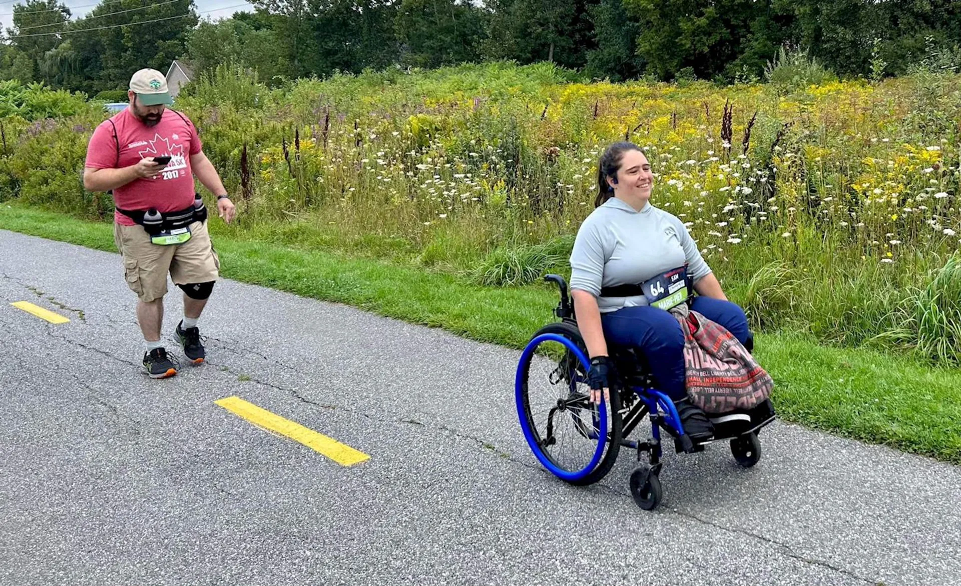 The image shows two people outdoors on a path lined with grass and wildflowers. One person is using a wheelchair, and the other is walking while checking a device in their hand. The setting appears to be along a nature trail with greenery and trees in the background.