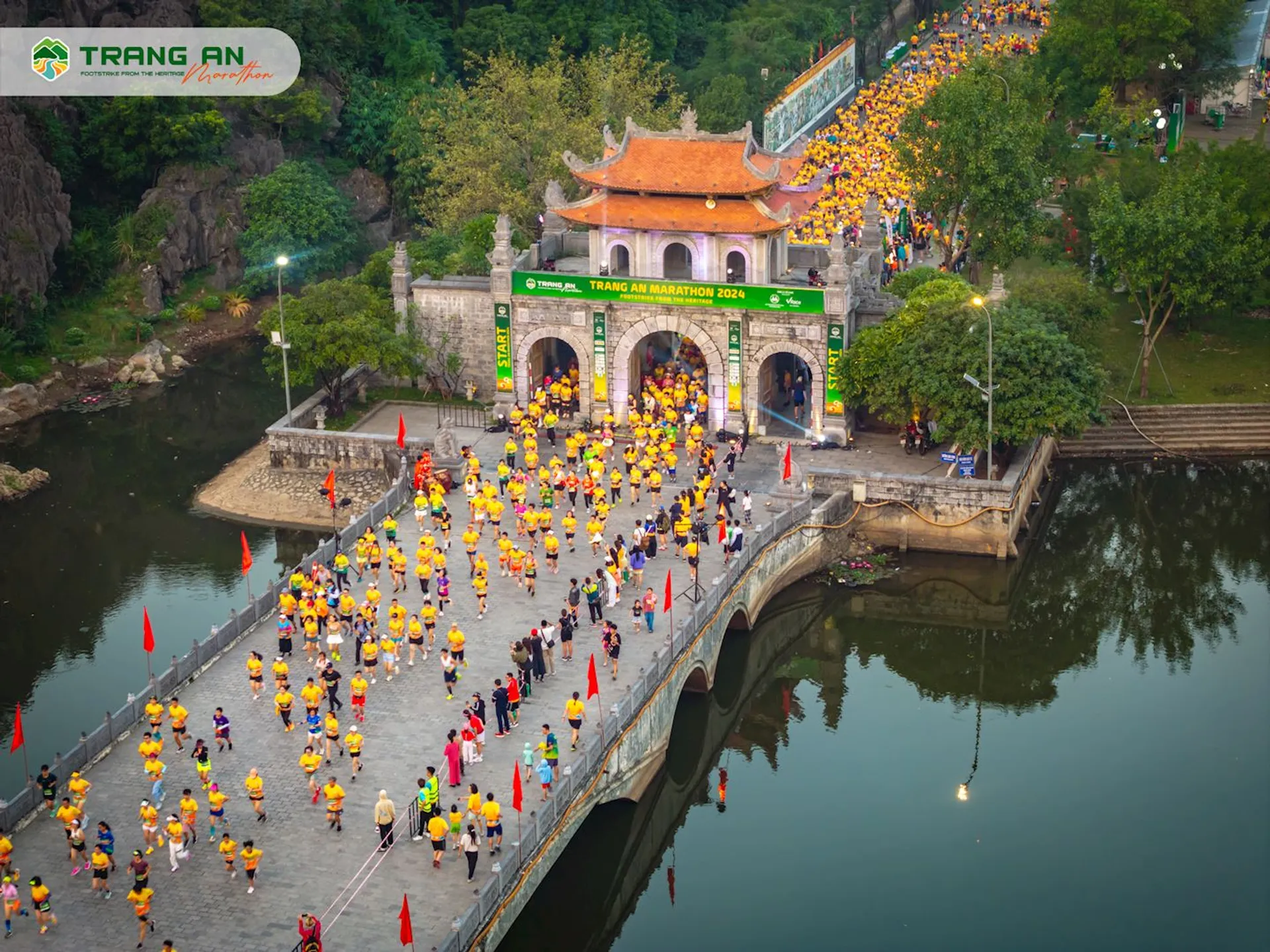 The image features a large group of people gathered on a bridge leading to a traditional gate-style entrance, likely for an event or celebration based on the coordinated clothing colors (yellow and orange tones). The structure has a distinctly East Asian architectural style with upturned eaves, which suggests that this could be a location in a country like Vietnam. There is lush greenery surrounding the area, with some trees and karst formations, which adds to the scenic beauty of the location. The banner at the top of the entrance spells out "TRANG AN," indicating that this is the Trang An Scenic Landscape Complex, a UNESCO World Heritage site in Ninh Binh province, Vietnam. Moreover, there are flags along the bridge and a crowd of people watching