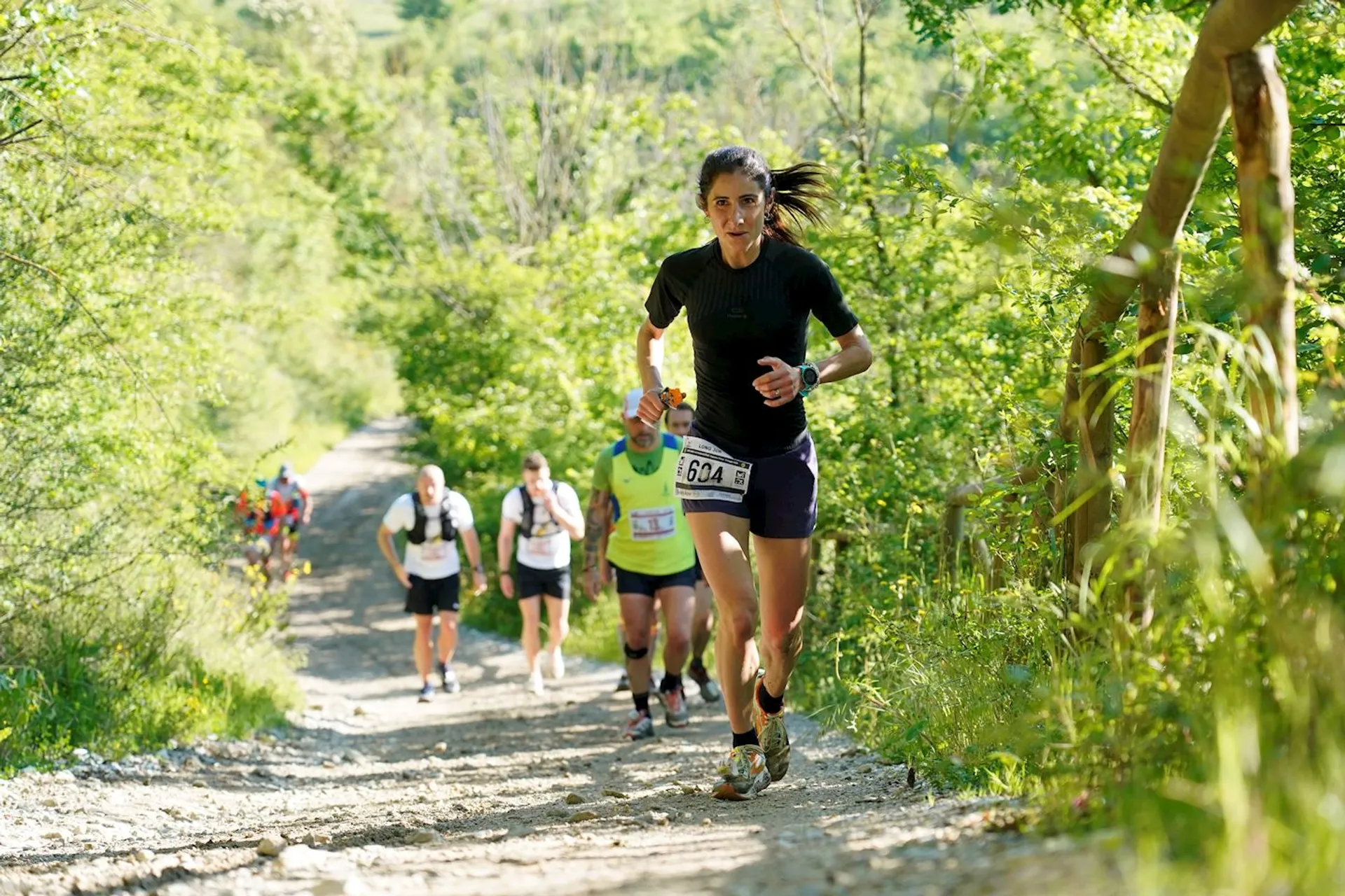 The image shows a group of people running on a dirt trail surrounded by lush greenery. They appear to be participating in an outdoor race or marathon. The trail is set in a natural environment with trees and bushes lining the path. The athletes are wearing running gear and race bibs.