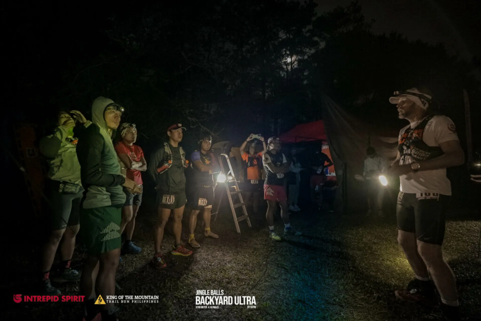 The image shows a group of people, likely runners, gathered outdoors at night, probably before a race event. They are standing with their attention directed towards a person on the right who appears to be addressing them. This might be a pre-race briefing or instructions being given. Headlamps and reflective clothing suggest that they are preparing for a night run or an event that extends into the night. The presence of signs like "INTREPID SPIRIT," "KING OF THE MOUNTAIN," and "BIG'S BACKYARD ULTRA" hints that this is an organized ultramarathon or endurance event. The setting appears to be a wooded or rural area with tents, indicating a base camp or staging area for the event.