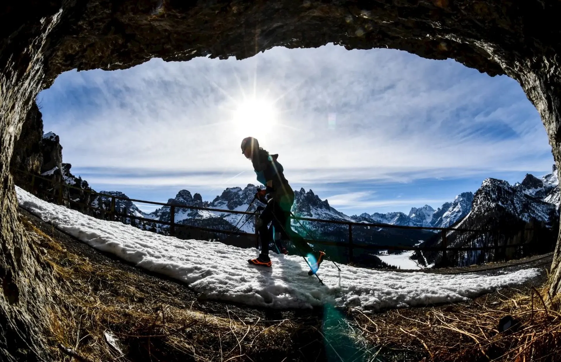 The image shows a silhouette of a person walking through what appears to be a tunnel or a natural archway. The person seems to be holding walking poles, which suggests that they might be hiking. The view through the opening frames a breathtaking mountain landscape under a stunning blue sky. The mountains are rugged and have snow-capped peaks, indicating this might be taken in a region known for alpine terrain. There's also snow on the ground, reinforcing the alpine or winter setting. The sun is shining brightly in the sky, creating a dramatic backlight effect. The photo is taken with a wide-angle lens, giving an expansive view of the scene and a feeling of openness. The fence to the side suggests the area is accessible and possibly a maintained hiking trail