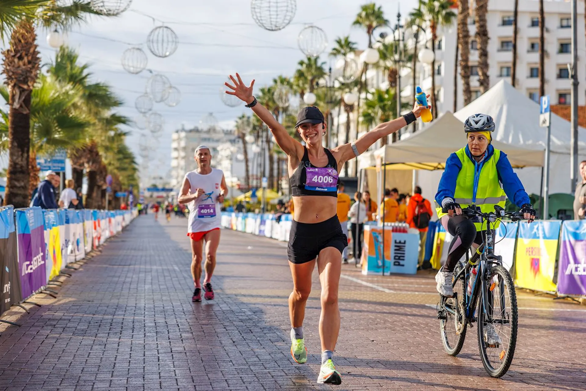 The image shows a running event on a paved street lined with palm trees. A female runner is in the foreground with her arms raised in celebration, wearing a sports bra and shorts with a participant number. A male runner follows behind her. To the right, a person on a bicycle is present, wearing a helmet and a yellow vest. Event tents and spectators are visible in the background. Decorative spherical lights hang above the street.