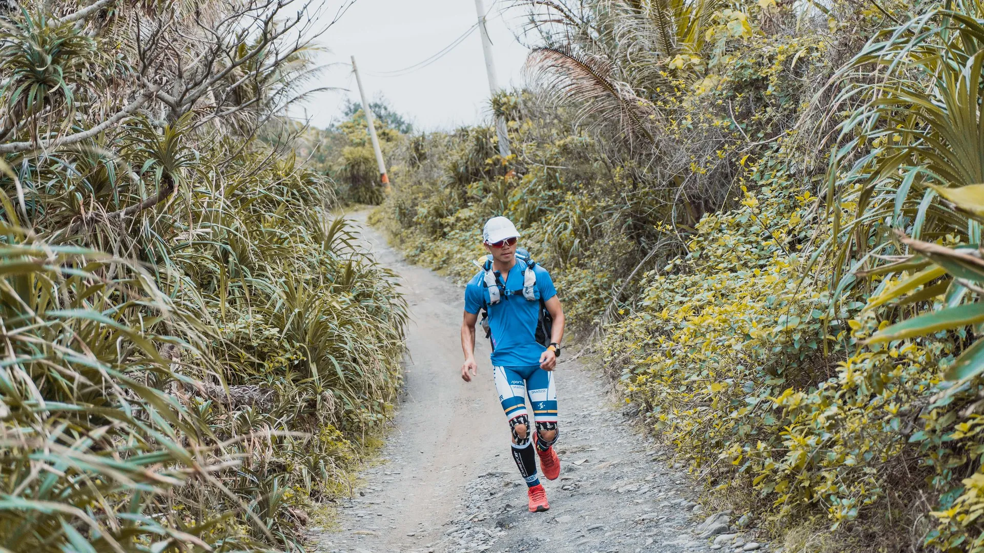 The image shows an individual jogging on a trail surrounded by greenery. The person is wearing a cap, sunglasses, a light-colored shirt, shorts, knee braces, and running shoes, and they are carrying a hydration pack on their back. The environment suggests a nature trail likely in a park or rural area. The weather appears to be fair, as the trail is dry and the person is dressed for warmer conditions.