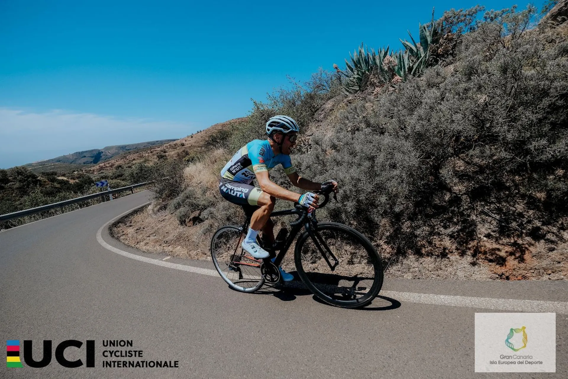 The image shows a cyclist riding on a road that twists through a dry, shrubby landscape. The cyclist is wearing a biking outfit with a helmet, sunglasses, and is on a road racing bike. The bottom of the image features the logos of the UCI (Union Cycliste Internationale) and a logo that appears to represent a cycling event—Granfondo Gran Canaria. This indicates that the photo is likely from a cycling competition or event on the island of Gran Canaria. The sky is clear, suggesting good weather conditions for cycling.