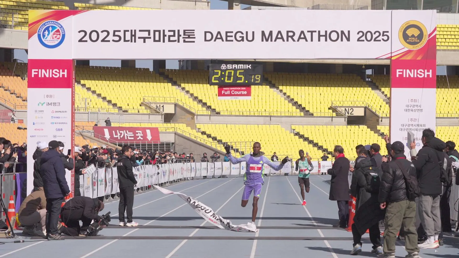 The image shows a runner breaking the finish line tape at the 2025 Daegu Marathon. The scene is set in a stadium, with spectators and photographers capturing the moment. A digital clock above the finish line displays the time as 2:05:19. The area is surrounded by banners and advertisements associated with the marathon event.
