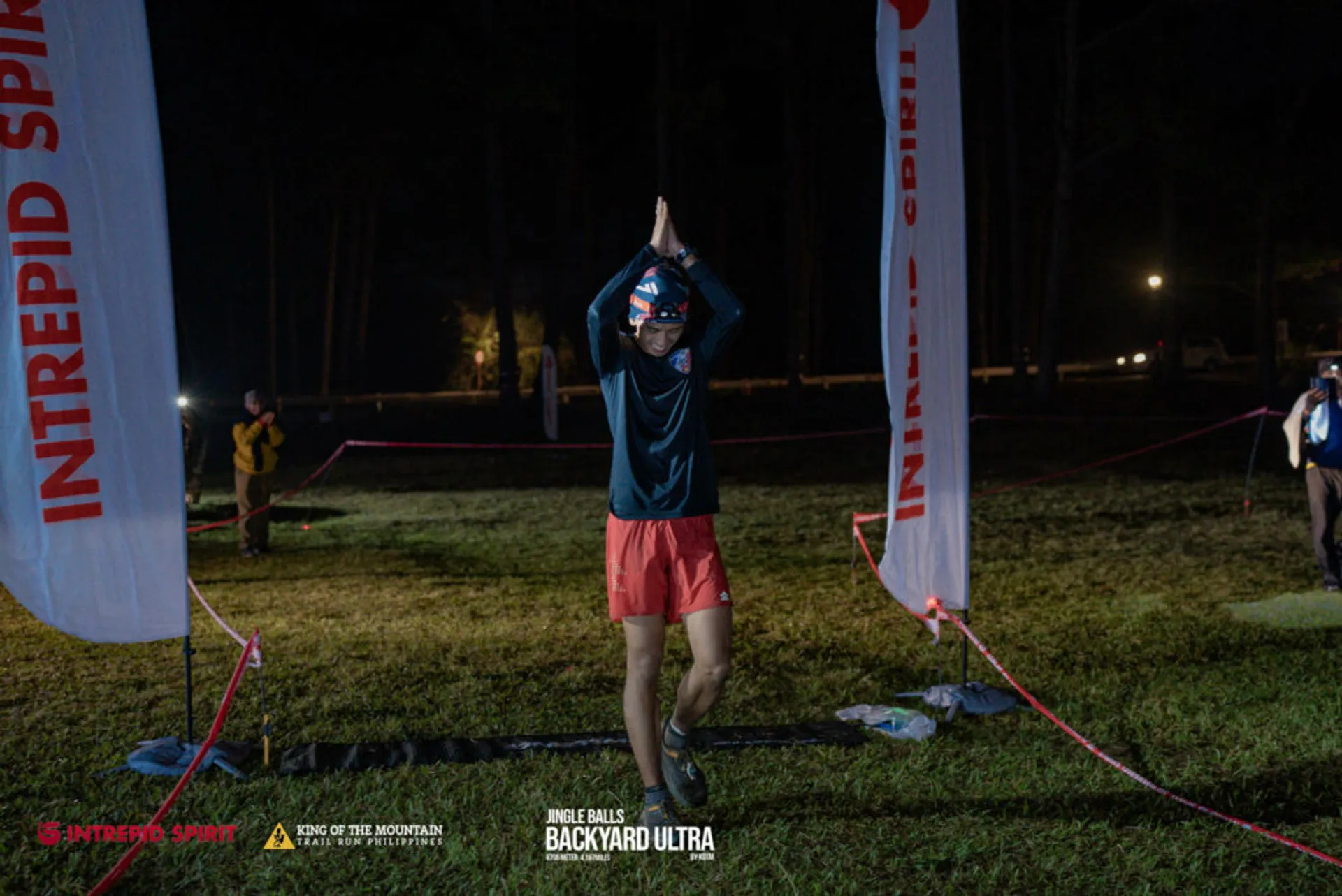 The image shows a person at night, likely a participant in an event called the "Intrepid Spirit Race the Mountain Backyard Ultra," as indicated by the banner in the background. This might be the finish line of a race where the participant is expressing joy or accomplishment by raising their arms up high, a common gesture when completing a challenge or celebrating a victory. The person is wearing a headlamp, a blue shirt, red shorts, and has a race number visible on their shirt, which is typical attire for an ultramarathon or long-distance trail race participant. In the background, other people, possibly race organizers, supporters, or fellow runners, can be observed as well as a forested area, suggesting that the event is taking place in an