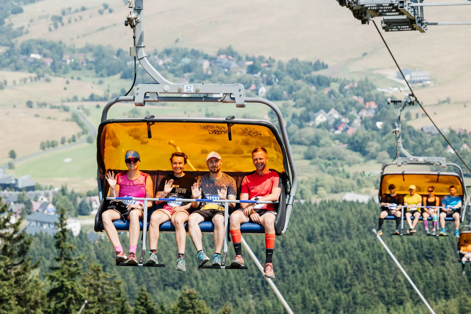 The image shows four people riding a ski lift chair, enjoying a scenic view. They are dressed casually, suggesting a summery or warm-weather setting. The background features a sprawling landscape with trees and hills, likely indicating a recreational or tourist area.