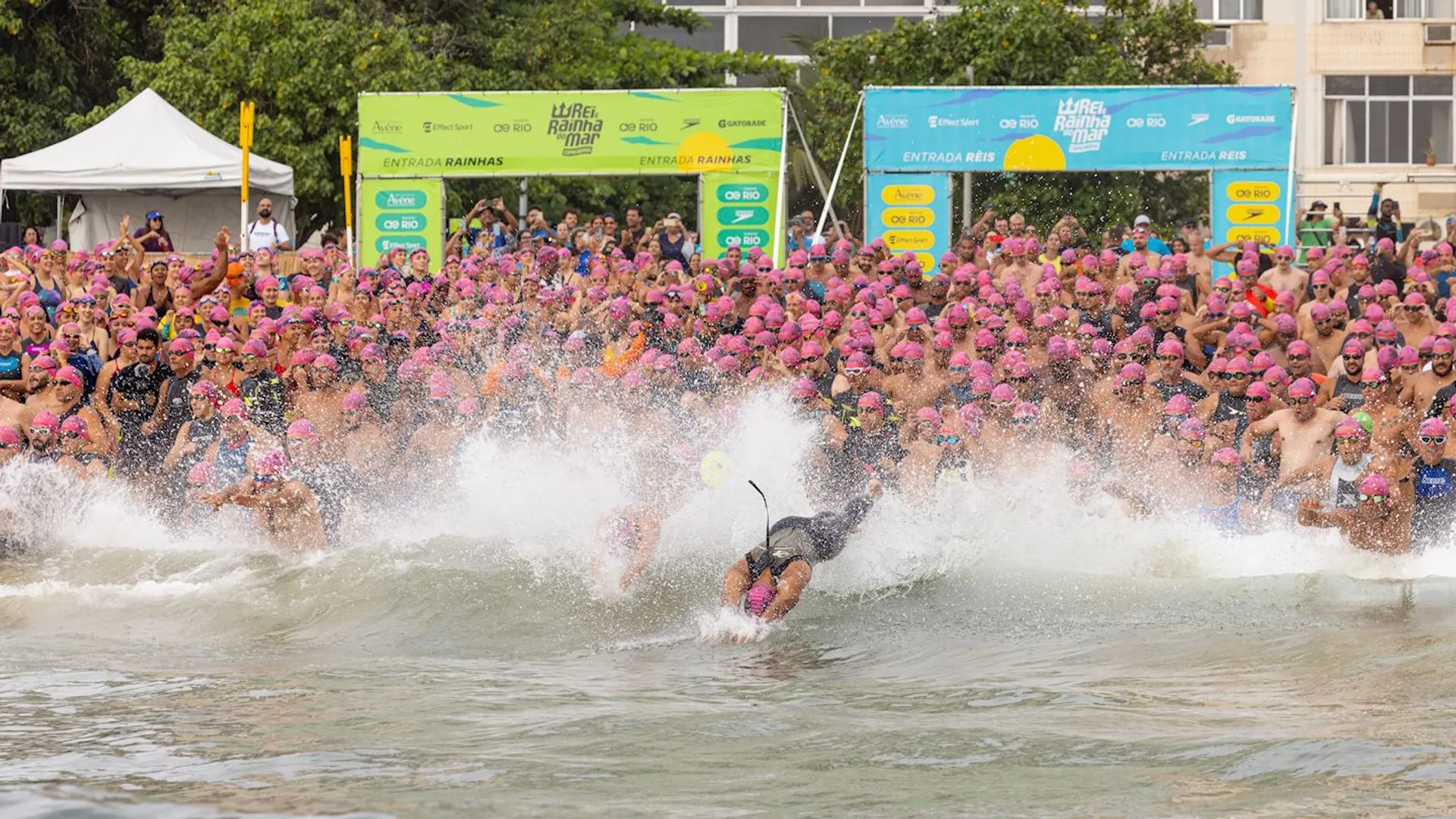 The image shows a large group of people wearing pink swim caps running into the water from the beach, likely participating in a swimming event or competition. There are banners in the background and a person diving into the water at the front of the group.