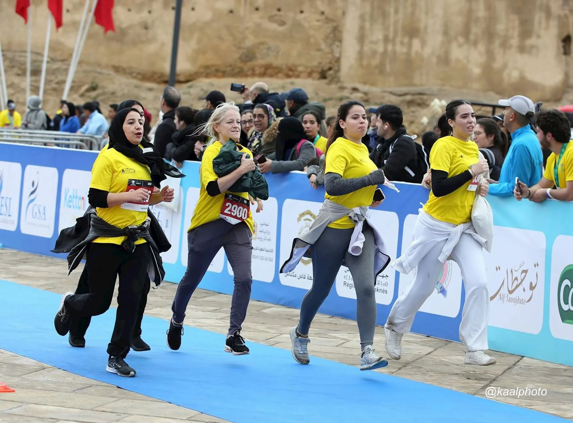 This image shows a group of women running in what appears to be a race or marathon. They're wearing yellow shirts and athletic clothing, some with jackets tied around their waists. There are spectators and other participants in the background, as well as signage and banners indicating it's an organized event.