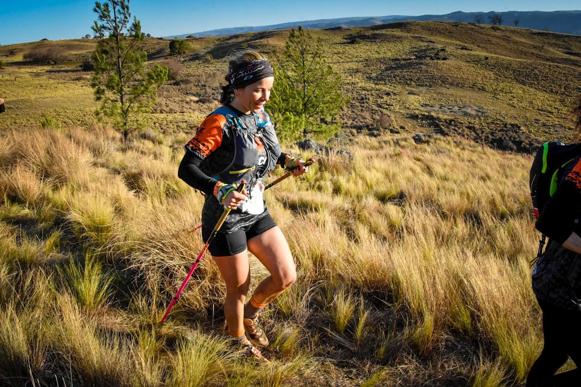 The image shows a person trail running through grassy terrain. They are equipped with trekking poles and wearing athletic gear, including a headband, a vest or hydration pack, and shorts. The landscape features hills and sparse vegetation, indicating a rugged and natural environment.