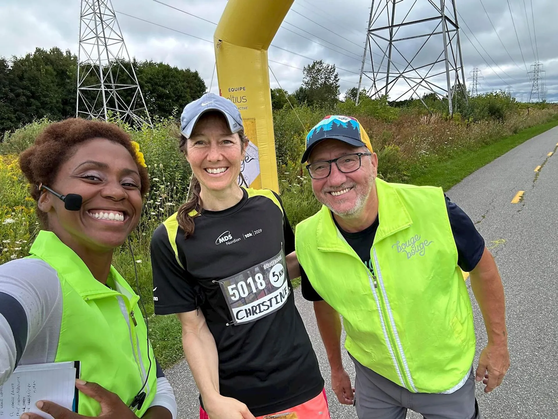 The image shows three people smiling for a selfie near a running or cycling path, likely at an outdoor event. One person is holding a paper, maybe for notes or timing. Another person is wearing a race bib with the name "Christine," suggesting they may be participating in a race or marathon. There are visible power lines in the background and some greenery along the path. They all appear to be in good spirits, possibly at a finish line or checkpoint.