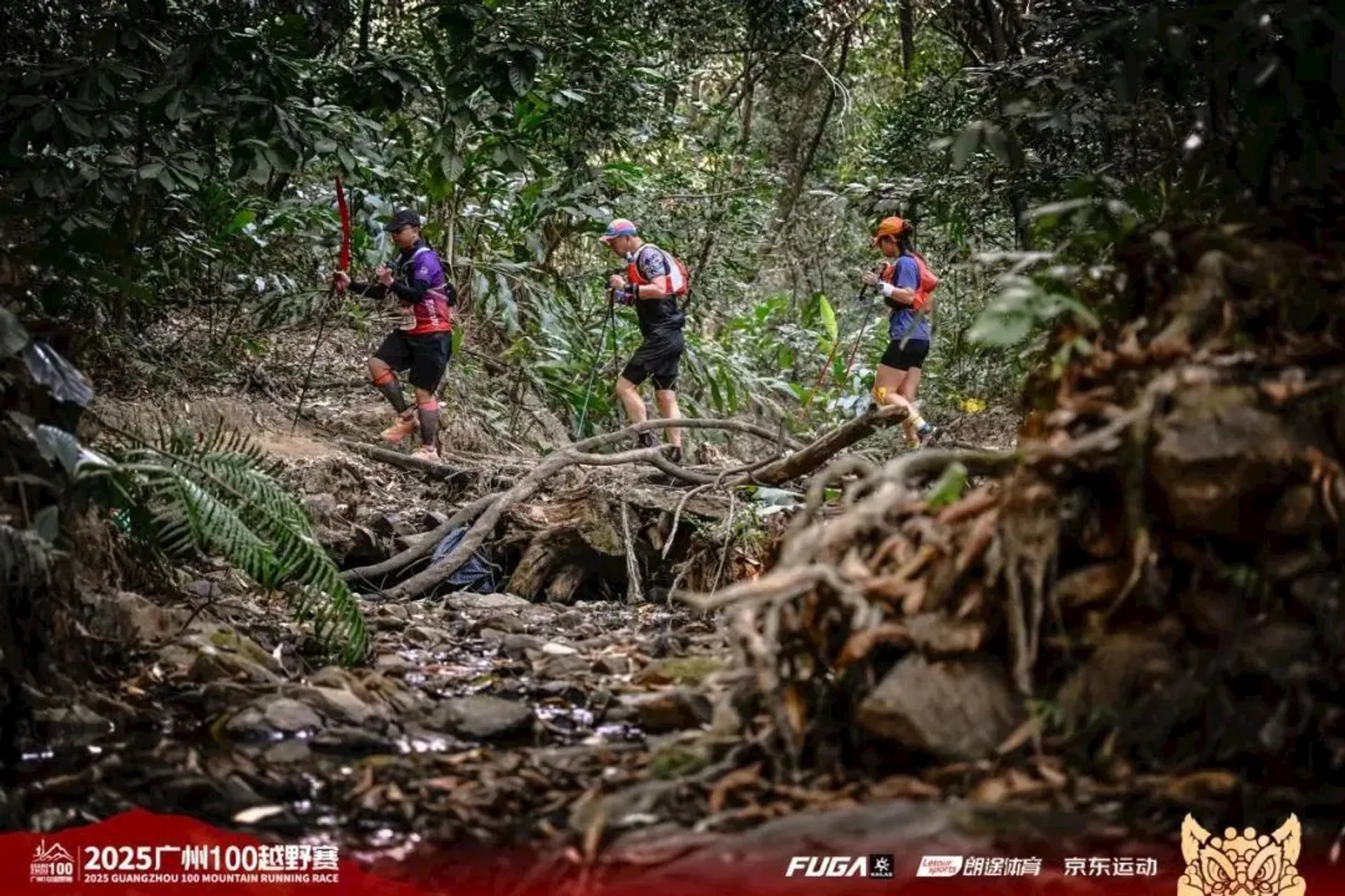 The image shows three people trail running through a forested area. They appear to be participating in a mountain running race, indicated by their gear and the organization logos. There is a rocky streambed in the foreground, and the scene is lush with greenery and trees. The event is labeled as the "2025 Guangzhou Mountain Running Race."