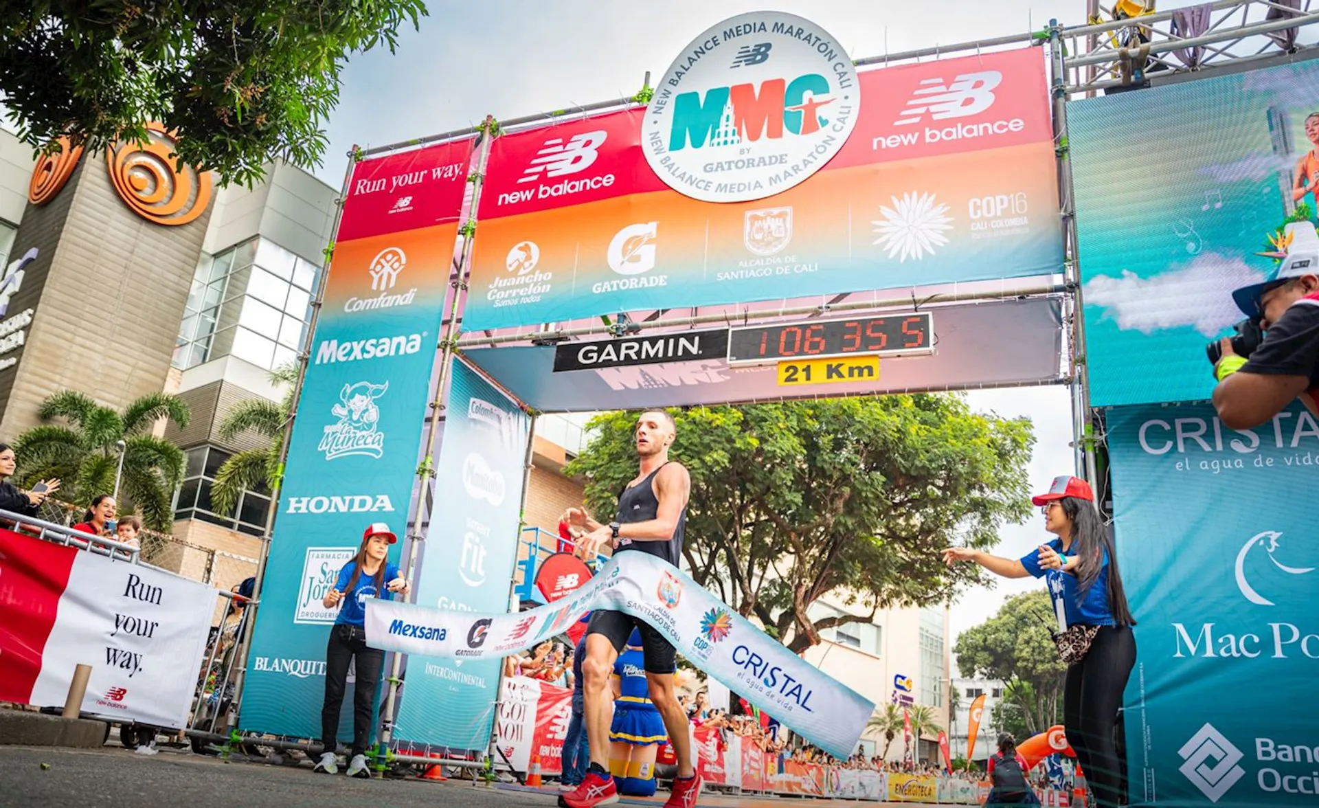 The image shows a runner finishing a half marathon at the New Balance Media Maratón. The finish line banner indicates it's a 21 km race. There are various sponsor logos visible on the banners, such as New Balance, Gatorade, and Mexsana. Spectators and staff are around the finish line, and there are buildings and trees in the background.