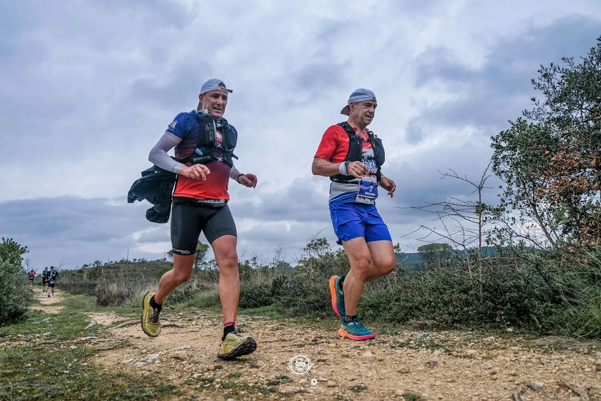 The image shows two people running on a dirt trail in an outdoor setting. They are wearing athletic clothing, including running shoes, and each has a hydration pack. The landscape appears natural with bushes and a cloudy sky. They look like they are participating in a trail race or run.