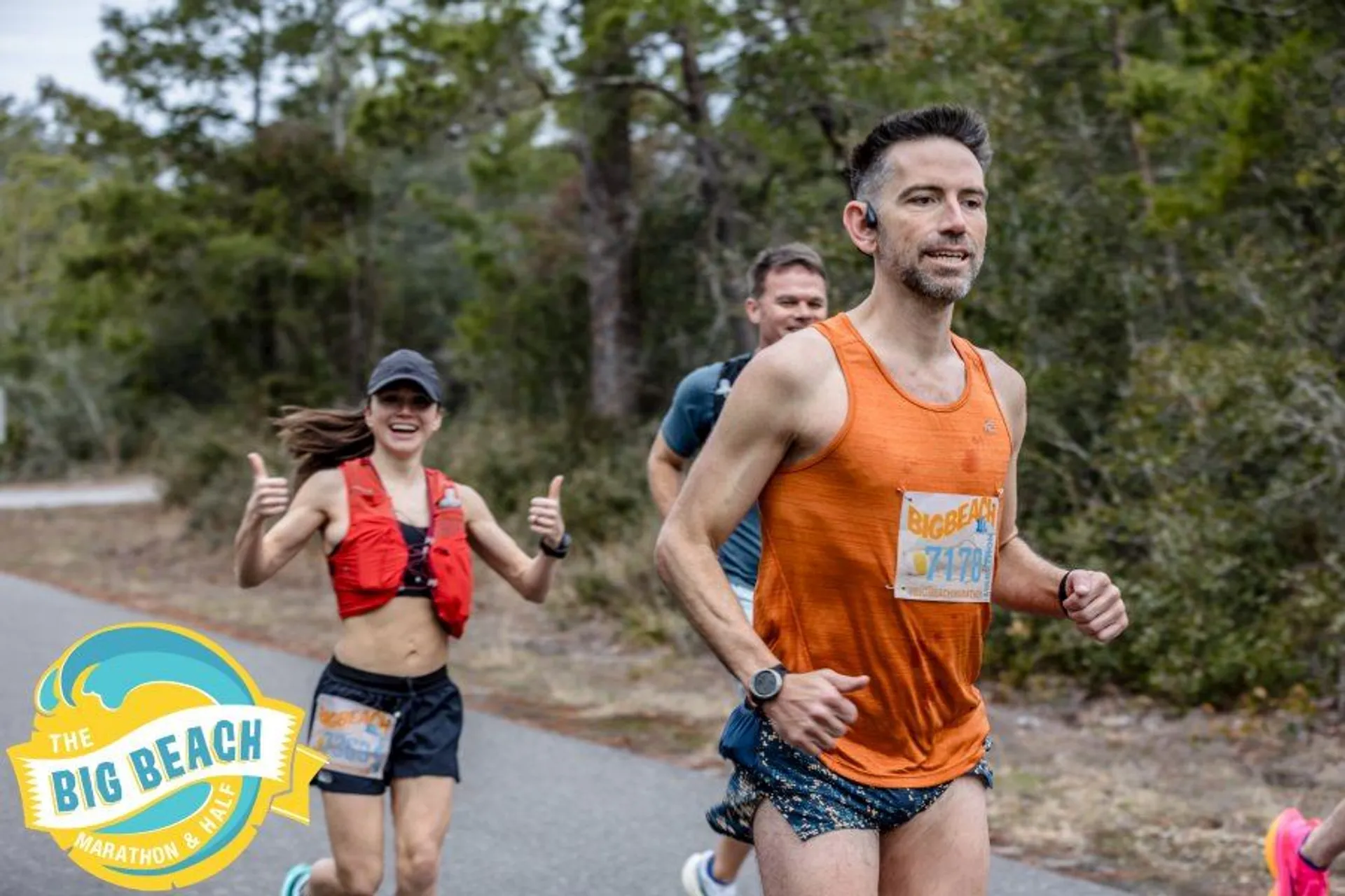 The image shows a group of people participating in "The Big Beach Marathon & Half Marathon." They are running along a road with trees in the background. One person is wearing an orange singlet, and another is wearing a red running vest and giving a thumbs-up gesture.