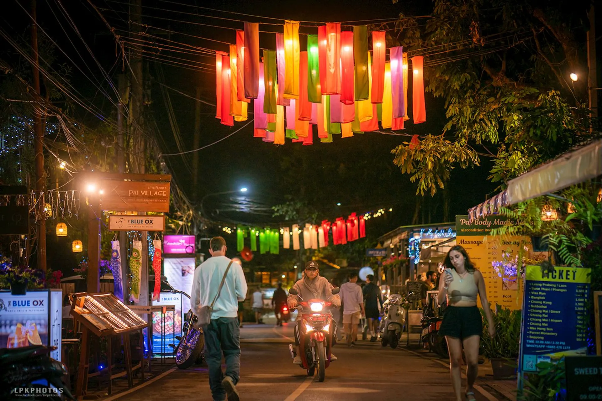 The image depicts a lively street scene at night featuring colorful hanging lanterns above the road. People are walking and riding motorcycles, and there are various establishments along the street with vibrant lighting and signs, indicating a bustling and festive atmosphere.