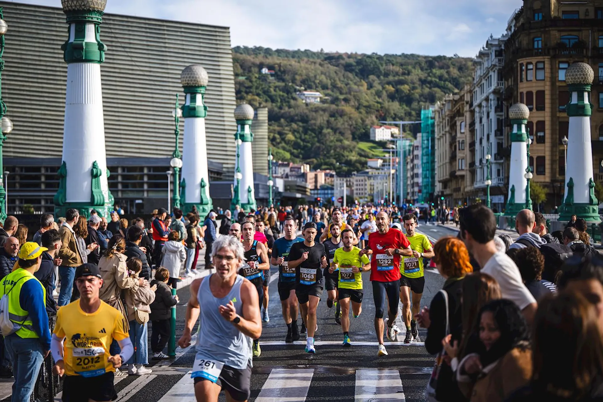 The image shows a marathon with many runners crossing a bridge. The street is lined with spectators observing the event. The background includes some modern buildings and a hillside with greenery. The scene appears to be vibrant and lively, capturing the activity and energy of the marathon.