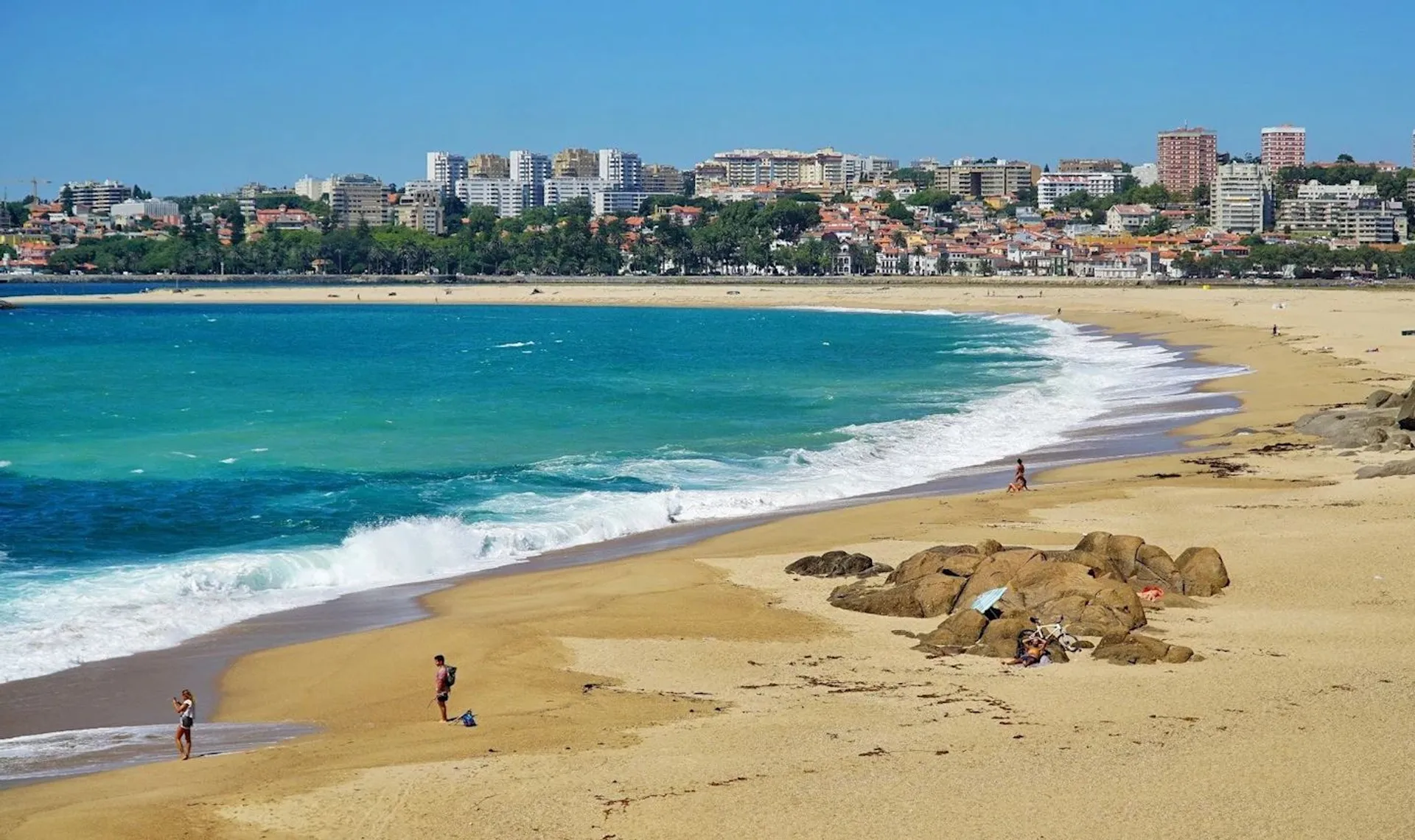 The image shows a sunny beach scene with clear blue skies. The shoreline curves gently with white waves lapping onto the golden sand. A few individuals can be seen near the water's edge, likely enjoying the beach activities such as walking or sitting on the sands. In the background, there is a cityscape with various buildings, including apartments or hotels, showing that this beach is adjacent to an urban area. The buildings appear to be built on a gentle rise, terraced up a hillside, and there are patches of greenery interspersed among the buildings. The sea is a vibrant shade of blue, suggesting it is a beautiful, clear day. Overall, it's a picturesque coastal scene likely to be a popular destination for recreation and relaxation.