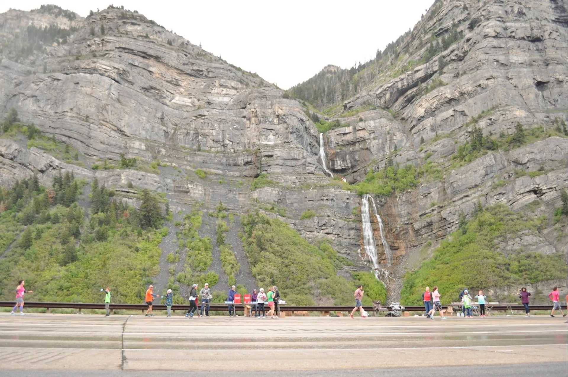 This image shows a scenic landscape with a large, rocky mountain in the background, featuring greenery and a waterfall cascading down the rocks. In the foreground, a group of people is walking or running along a road, suggesting an outdoor event or activity, such as a hike or marathon, near the mountains.