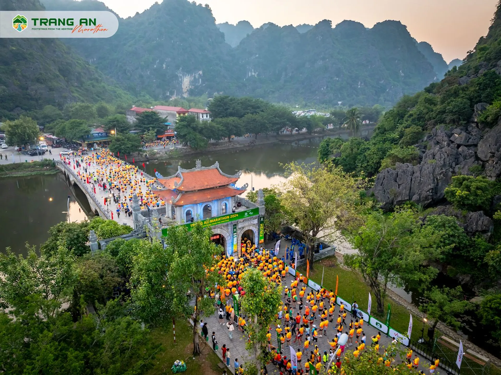 The image shows a lively cultural or religious event taking place in a scenic location. A large group of people, many of whom are carrying yellow and orange lanterns or balloons, are walking along a path that leads to a traditional archway or gateway. Beyond the gate, there is a bridge over a body of water that connects to a temple or pagoda complex with red roofs, which is set against a backdrop of dramatic karst limestone mountains.

The landscape suggests that this might be taking place in a region known for its natural beauty and possibly of cultural or spiritual significance, such as Trang An in Ninh Binh Province, Vietnam (as indicated by the watermark "Trang An Festival"). The tranquil water, lush greenery, and towering mountains