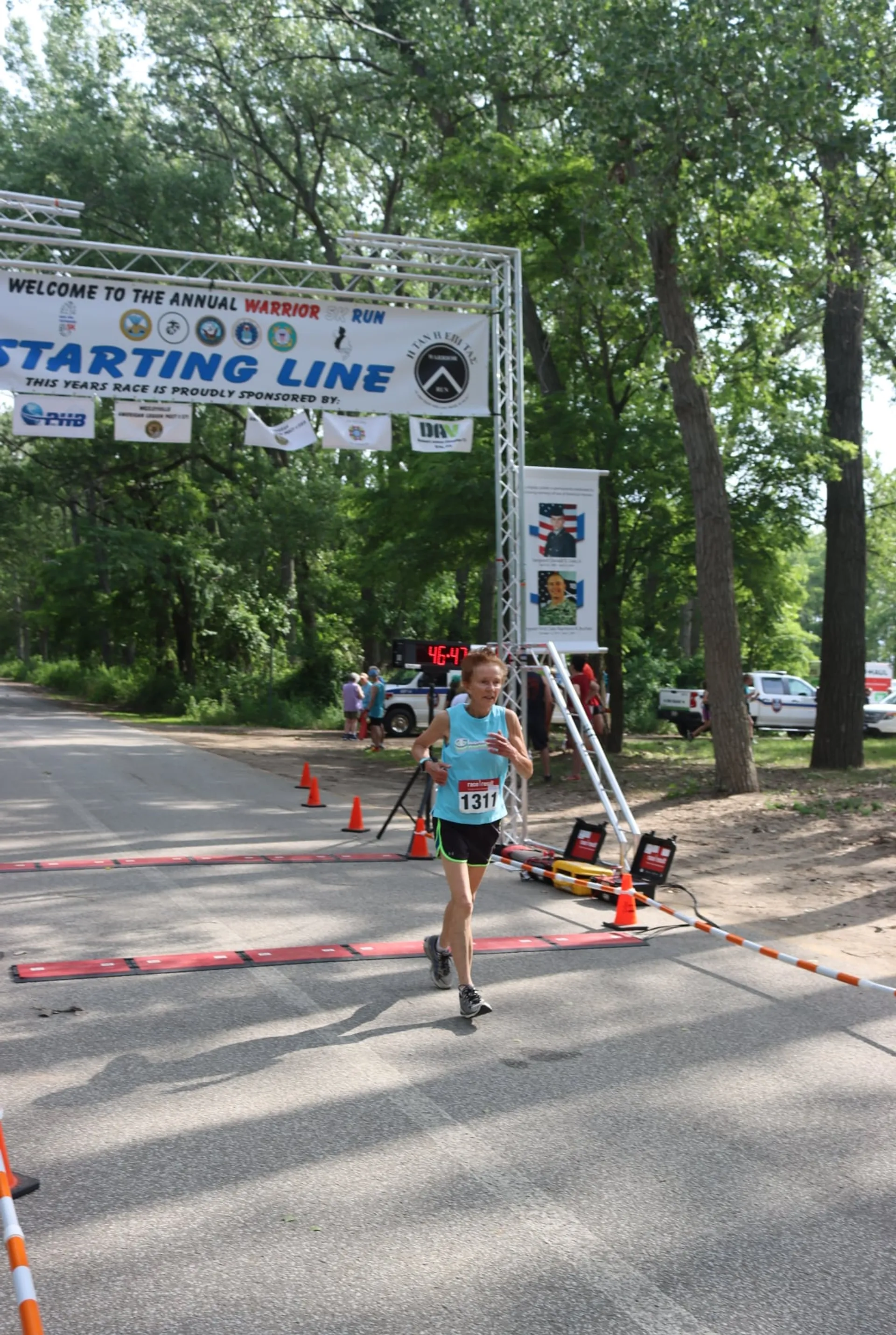 This image shows a young athlete crossing the finish line of a running event. The banner above denotes this as the "Starting Line" of the annual Warrior Run, but it appears to be used also as a finish line here. The runner is wearing a race bib with the number "1317" and appears to have just completed the race, based on their solo position and the relaxed posture after likely pushing through to the end. There are cones marking the end of the race path, a timing device to the side, and some spectators in the background. The event is set in a park-like setting with trees and greenery around, and it's a sunny day which suggests good weather for the event.