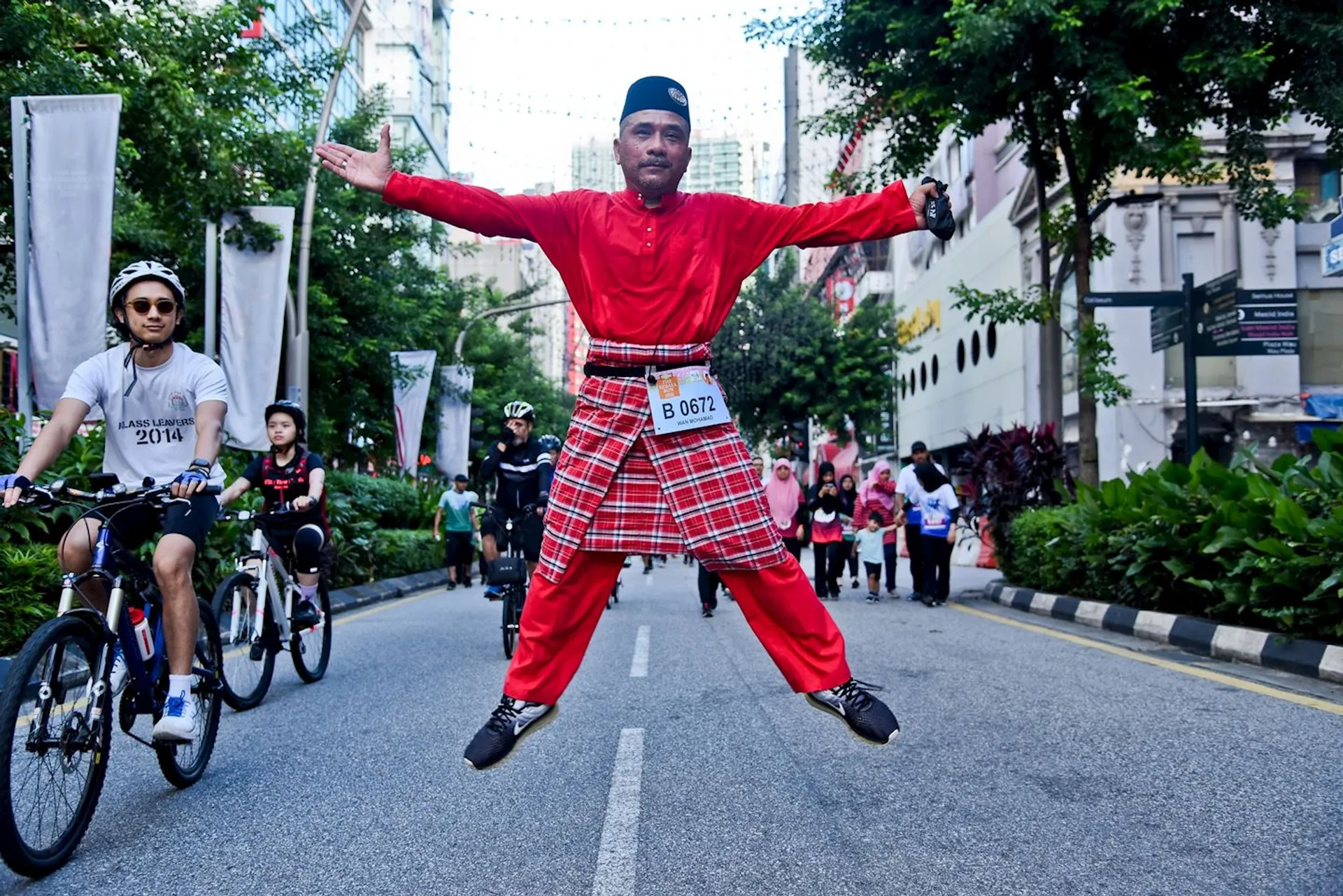 This image shows a person wearing traditional attire jumping energetically in the middle of a street during an event. The person is surrounded by people on bicycles. The street is lined with trees and banners, and there are buildings in the background.