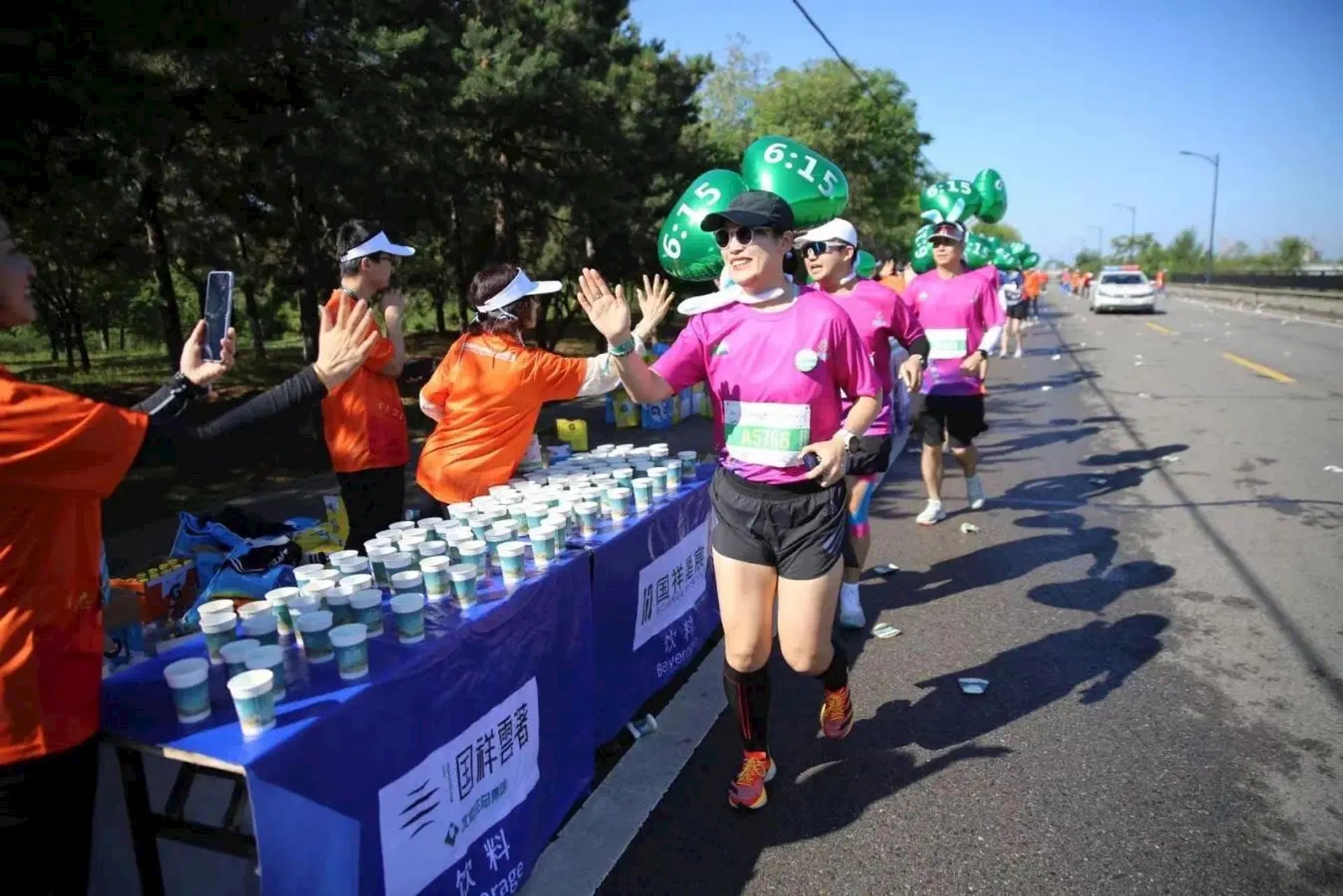 The image shows a group of runners participating in what seems to be a marathon or running event. There are tables with hydration stations providing drinks. The runners are wearing race bibs, and some have large balloons with numbers like "6:15" attached to them, likely indicating pace groups. The event staff are wearing orange shirts, and the setting appears to be outdoors with many trees in the background.