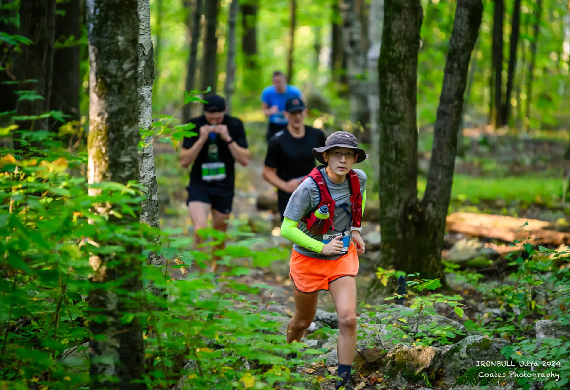 The image shows a group of people trail running through a forested area. The main person in the foreground is wearing a hat, a vest, an orange skirt or shorts, and running shoes. Other runners are visible in the background, surrounded by green trees and foliage. The setting suggests it's part of an outdoor race or an organized running event in a natural environment.