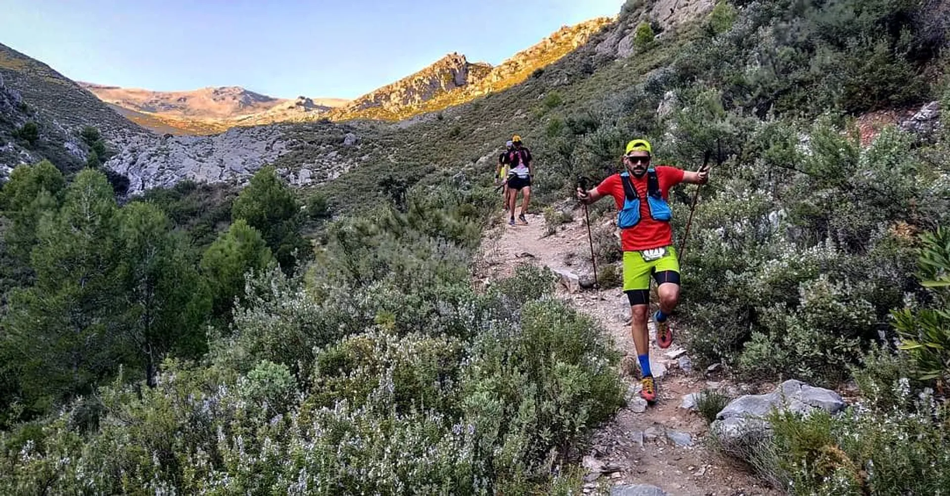 The image shows two individuals participating in trail running or hiking in a mountainous outdoor setting. The foreground features a runner wearing a bright yellow cap, sunglasses, a blue shirt with red accents, black shorts, and running shoes. Additionally, the runner is equipped with a hydration pack on their back. The background runner is similarly geared for an active outdoor adventure, wearing a hat, a hydration pack, and appropriate attire for the activity. They appear to be following a narrow dirt trail surrounded by natural vegetation typical of a rugged terrain, and the time of day looks to be either early morning or late afternoon, as suggested by the long shadows and the warm lighting. Mountains loom in the distance, completing this picturesque scene.