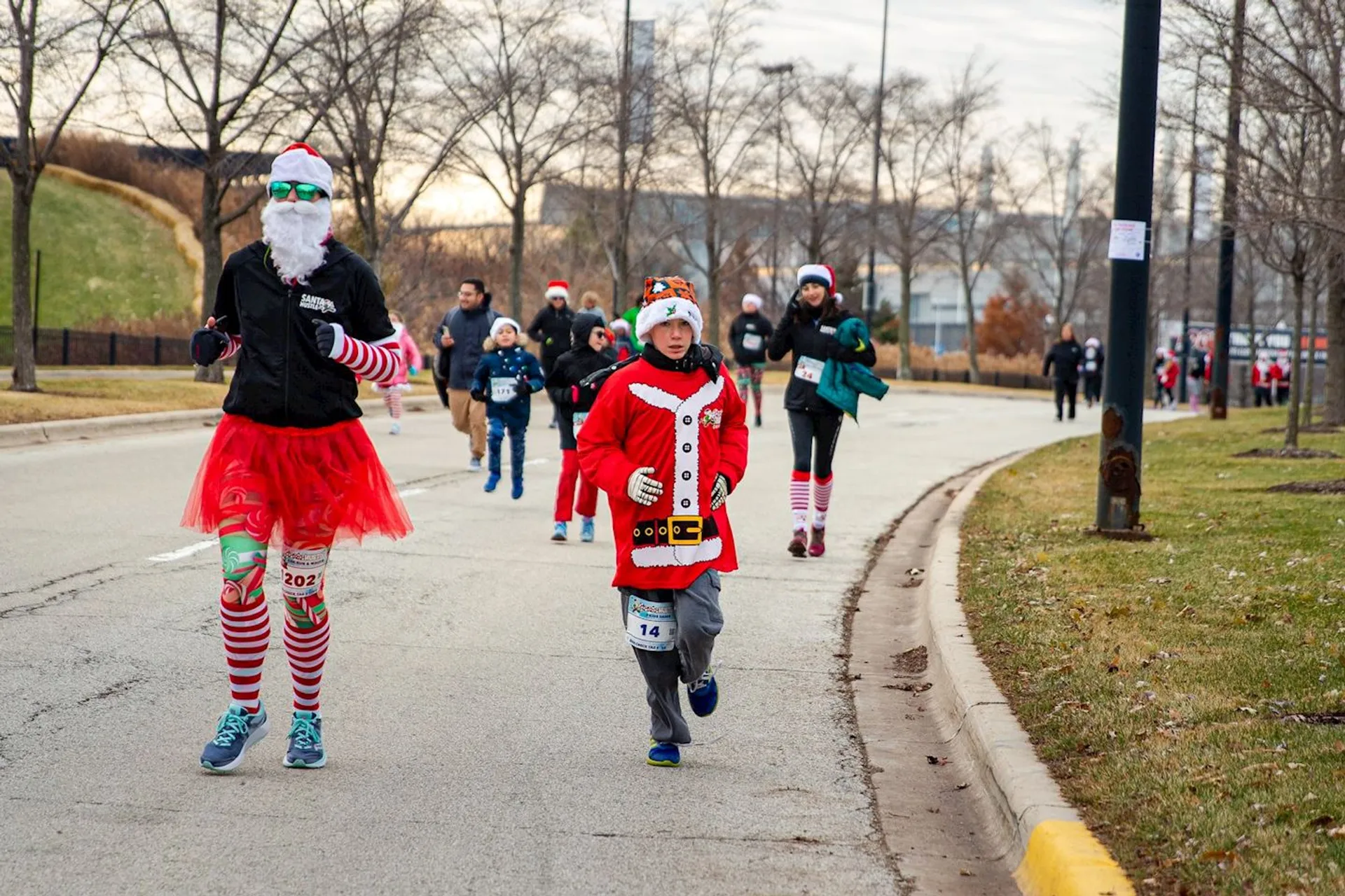 The image shows people participating in a festive run. Some individuals are dressed in holiday-themed costumes, including outfits resembling Santa Claus. The atmosphere appears cheerful, with participants jogging along a paved road lined with trees.