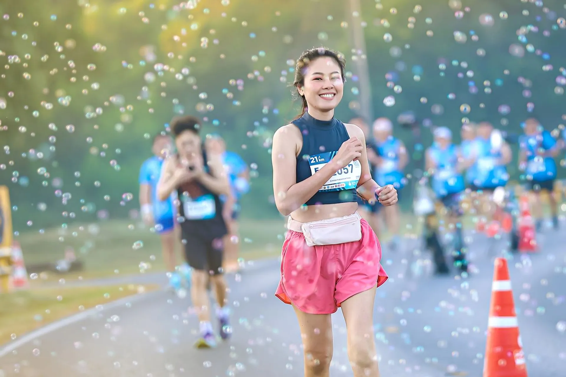 The image shows a person running in a race with a joyful expression. They are wearing a bib number and surrounded by bubbles. There are other runners and orange cones visible in the background, indicating a race event.
