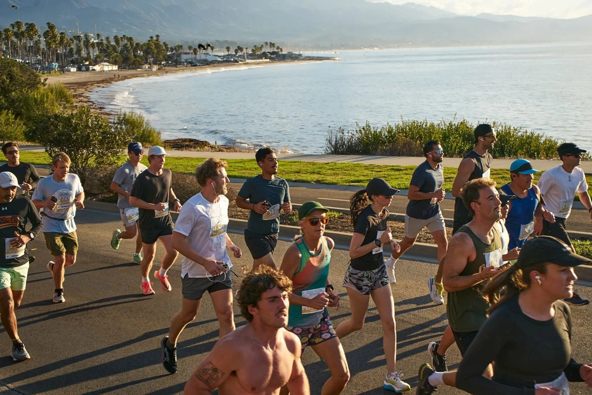 This image shows a group of people running along a coastal path. The background features a scenic view of the ocean, palm trees, and mountains. The runners are wearing athletic gear and appear to be participating in an organized event, such as a race. The setting suggests a pleasant day with clear skies.