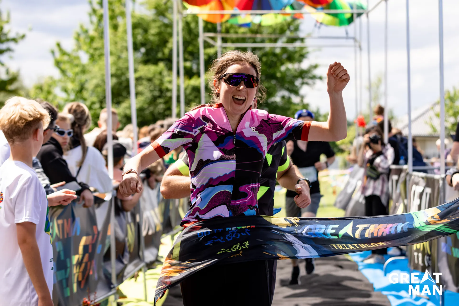 The image shows a person crossing the finish line of a race, likely a triathlon, as indicated by the text on the banner and the outfit. The person appears to be celebrating their achievement. There are spectators in the background, and the event seems lively, with bright colors and a joyful atmosphere.