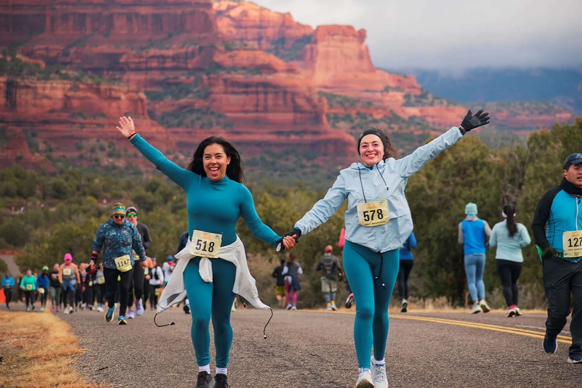 The image shows two people running a race in an outdoor setting with mountainous terrain in the background. They are wearing running gear and have bib numbers pinned on their clothing. The scene appears lively with more participants in the background, suggesting it's part of a larger event. The landscape features red rock formations.