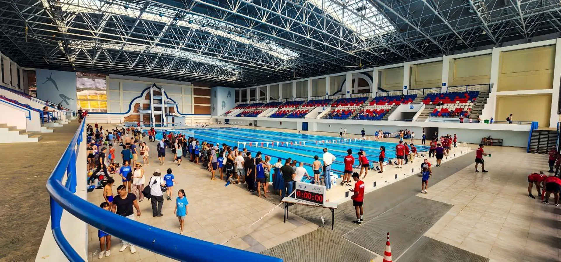 The image shows an indoor swimming pool facility with a large pool in the center. There are many people around, including swimmers and spectators. The seating area is filled with people, and it looks like a swimming event or competition is taking place. The architecture includes a high ceiling with a lot of natural light coming through windows.