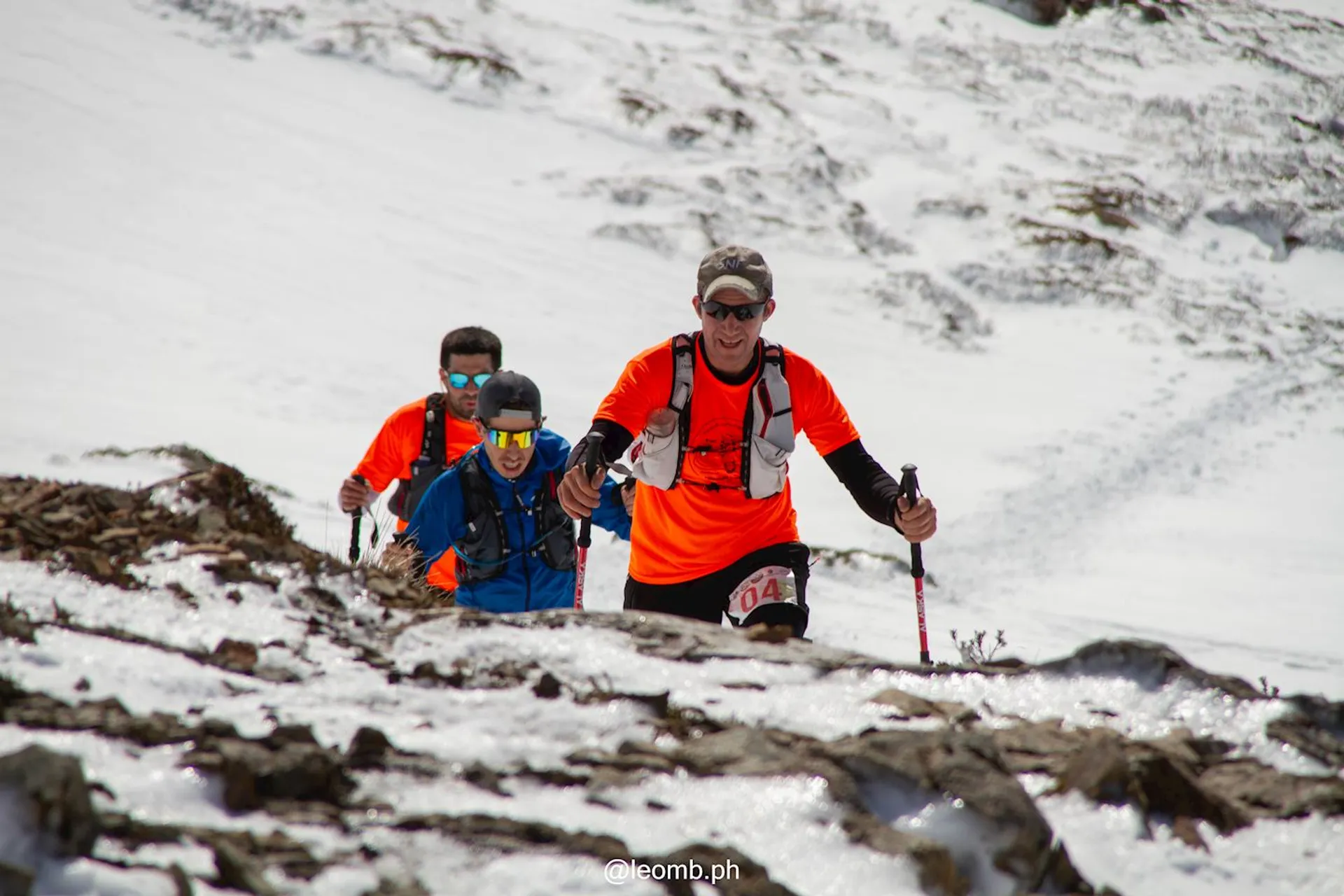 The image shows a group of people hiking or trekking in a snowy mountainous area. They are dressed in bright clothing and appear to be engaged in an outdoor adventure, using hiking poles to assist their movement through the snow.